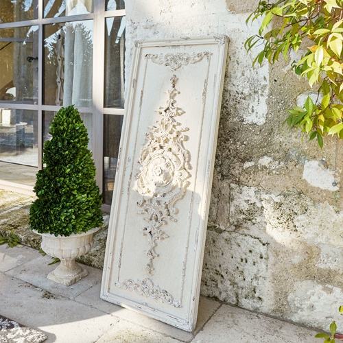 Pannello decorativo in legno bianco con rilievo vintage appoggiato a muro di pietra accanto a pianta verde