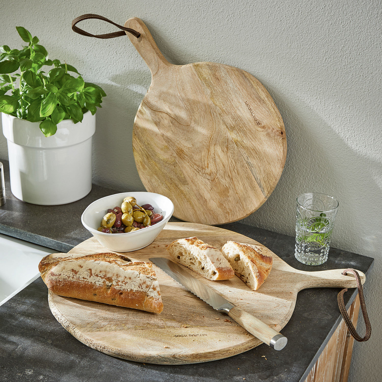 Coppia di taglieri Nitra Tagliere rotondo in legno naturale con manico, coltello da pane, pane affettato e olive su piano cucina