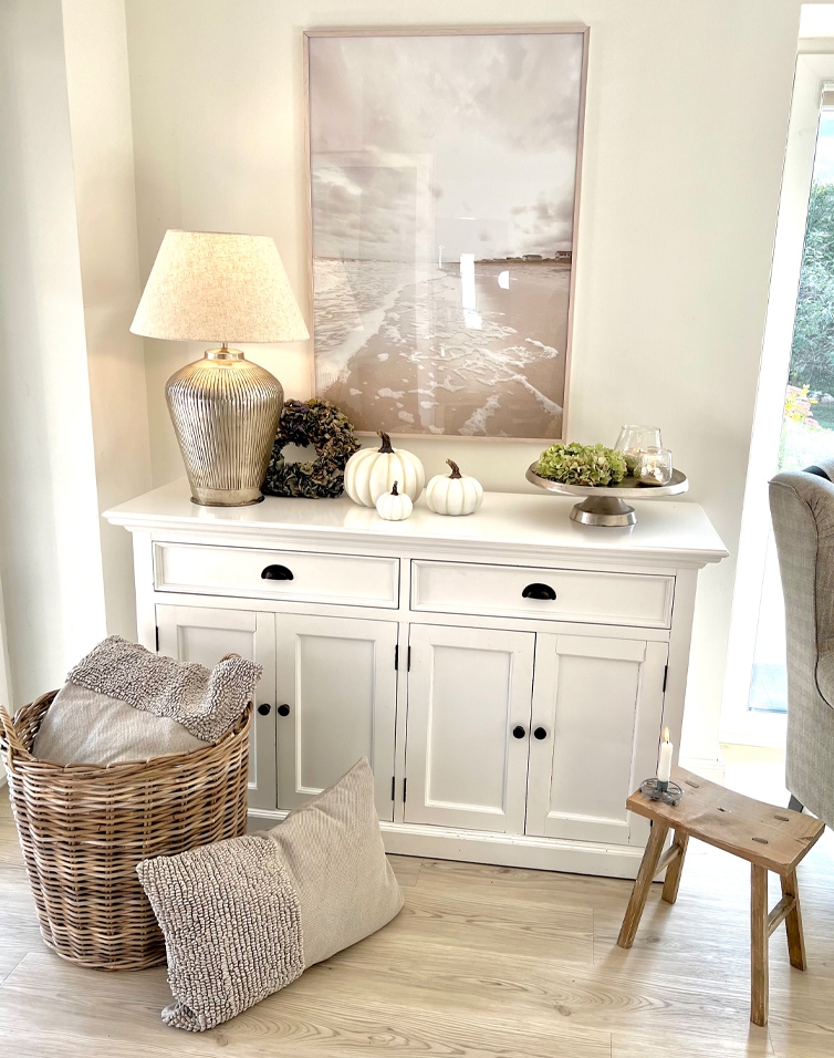 Madia bianca in legno con lampada, zucche decorative e cesta di vimini con cuscini in soggiorno luminoso