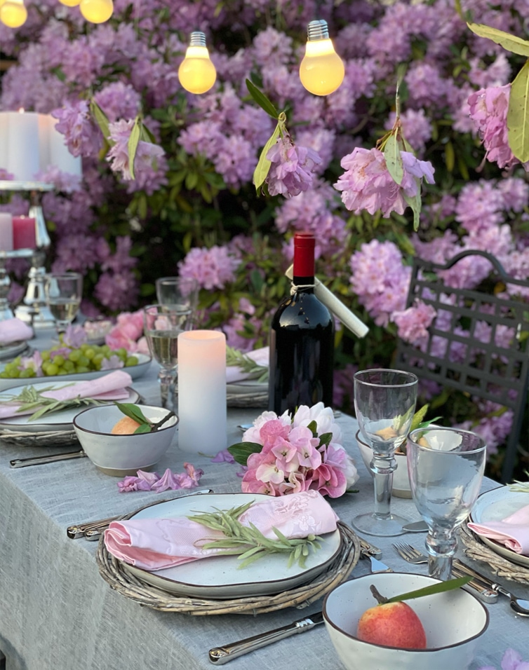 Allestimento estivo con fiori rosa, luci e bottiglia di vino davanti a cespugli viola.