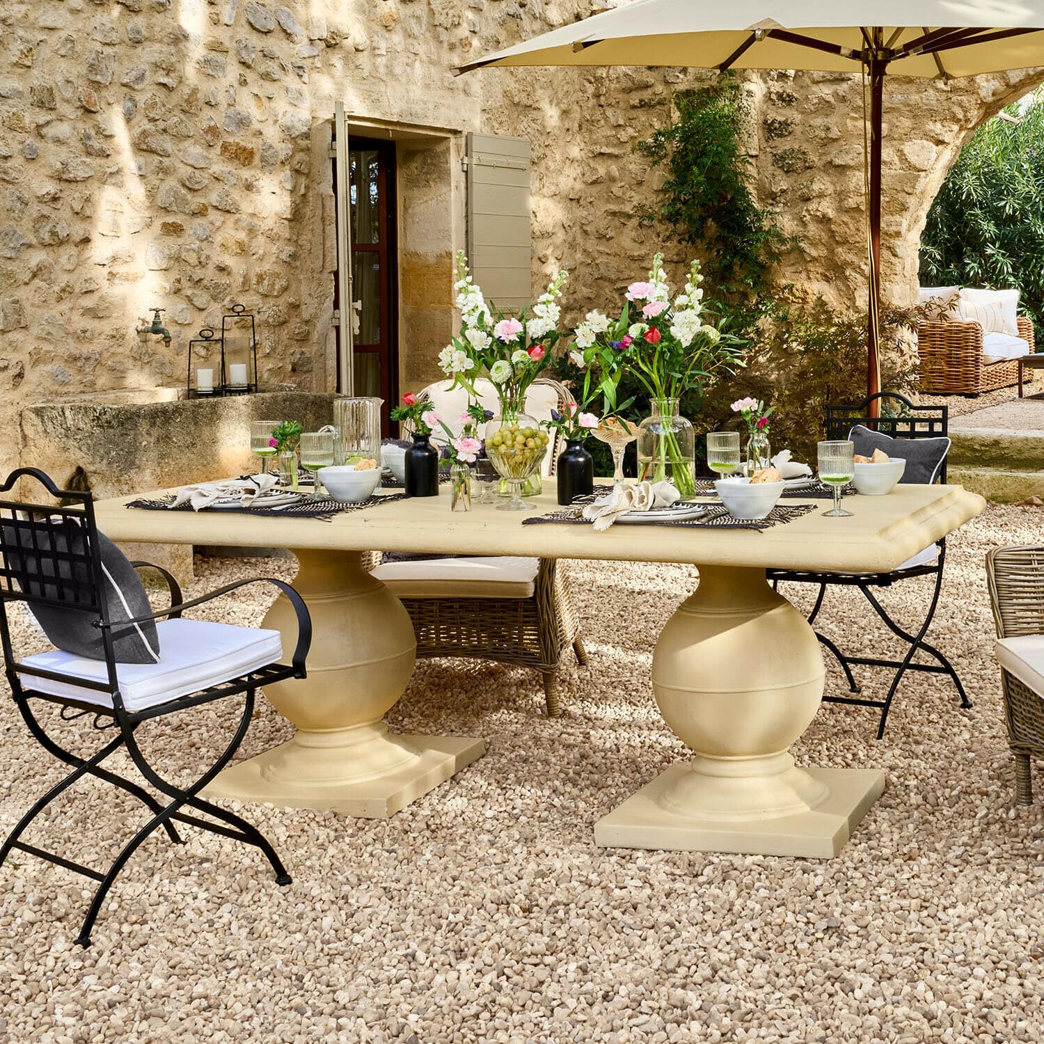 Tavolo Talvero Grande tavolo da giardino beige in pietra su patio ghiaioso con fiori e sedie in metallo e vimini
