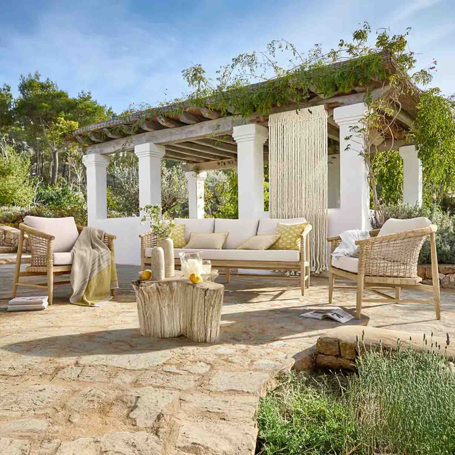Set di mobili da giardino in legno chiaro con cuscini crema su terrazza in pietra sotto pergolato con piante