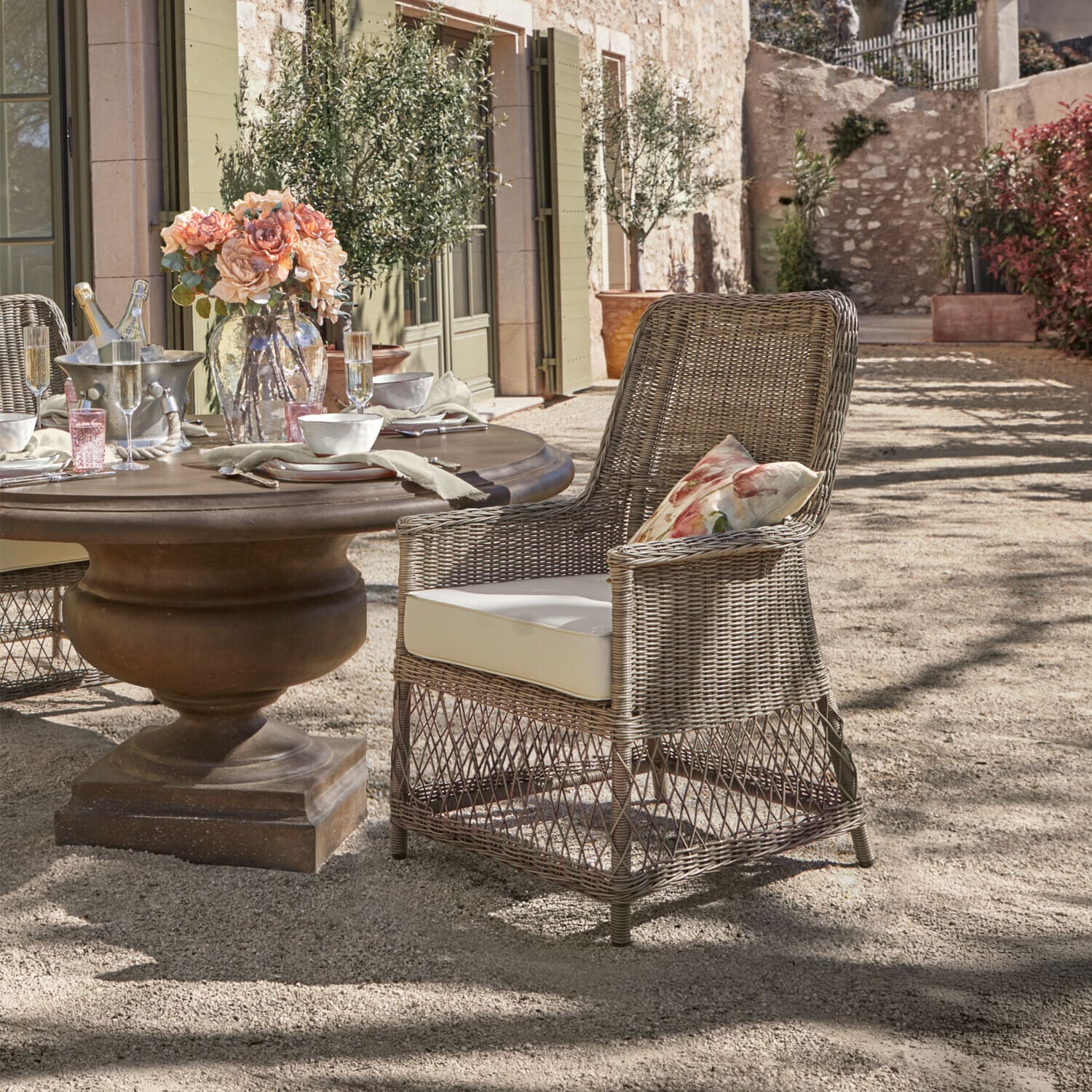 Poltrona Fenris Sedia in rattan grigia con cuscino crema e cuscino floreale accanto a tavolo rotondo all'aperto