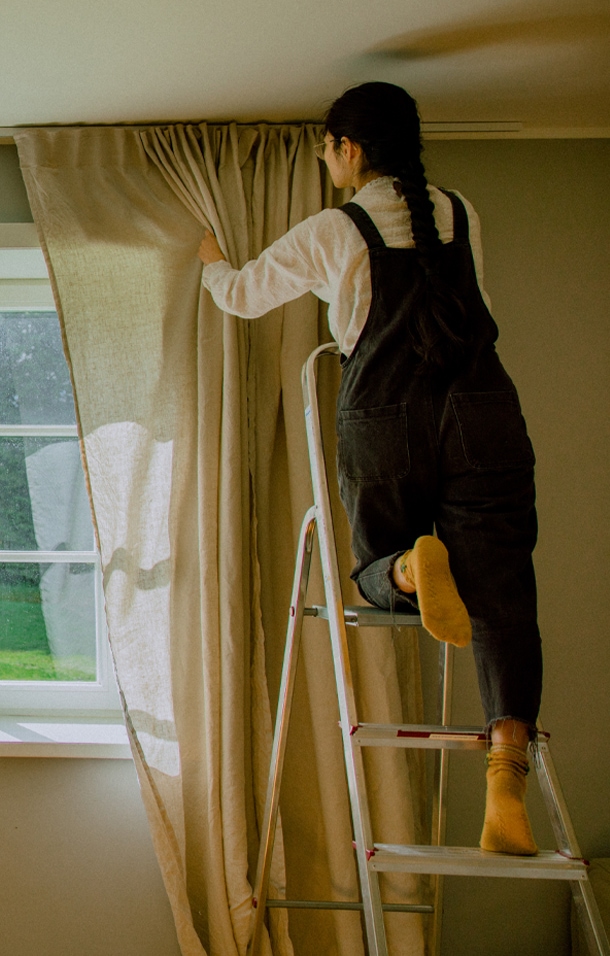 Donna su scala che sistema una tenda beige in lino davanti a una finestra in una stanza luminosa