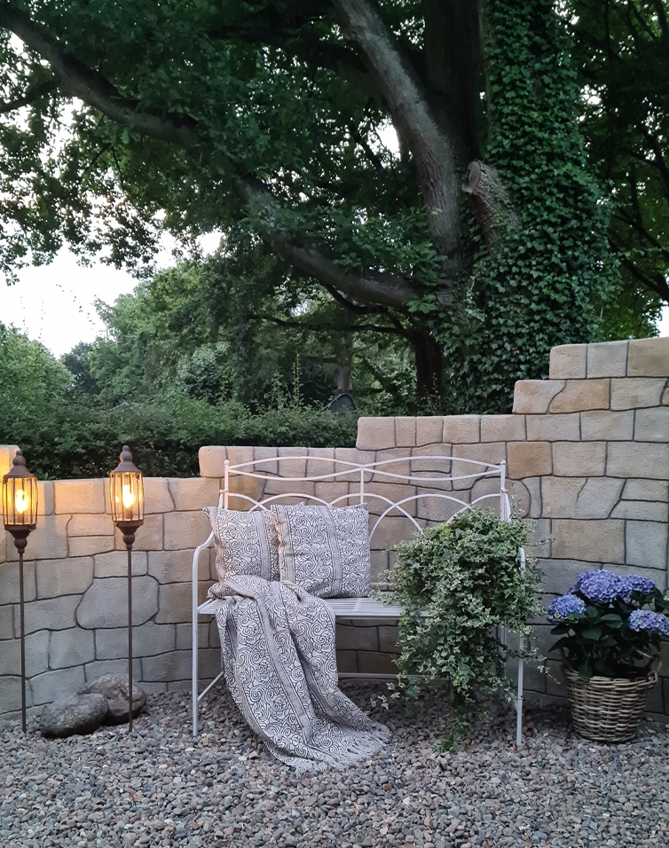 Panchina da giardino in metallo bianco con cuscini e coperta, circondata da muro in pietra e piante
