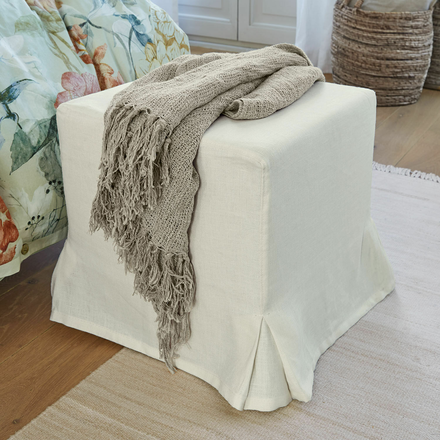 Pouf Ulysette Pouf beige in tessuto con coperta lavorata a maglia grigia su pavimento in legno in ambiente accogliente