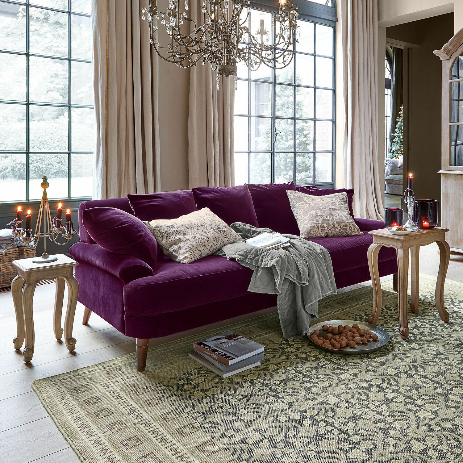 Divano Rivermere Divano in velluto viola con cuscini e coperta grigia in soggiorno luminoso con grandi finestre e tavoli in legno