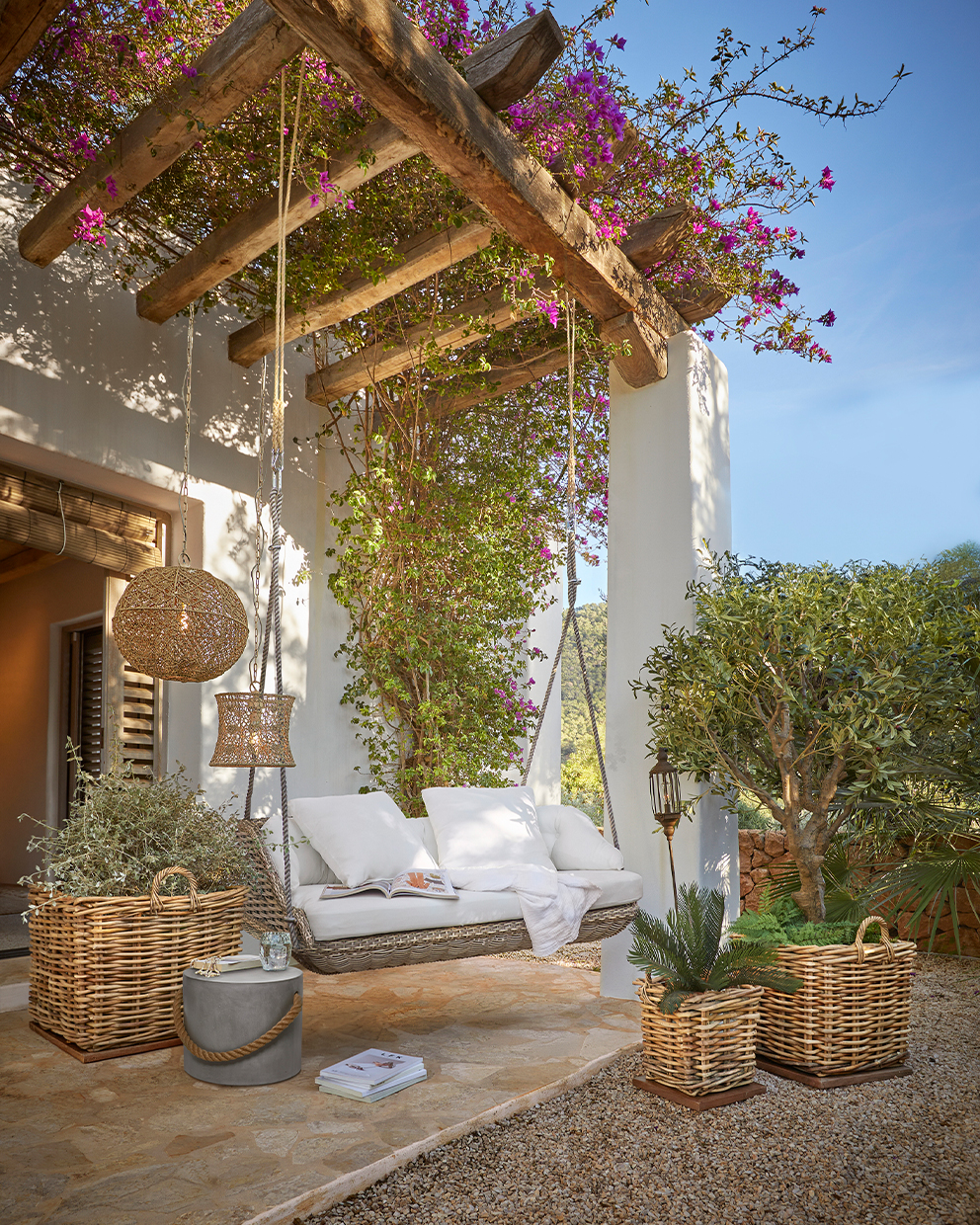 Dondolo da giardino in vimini con cuscini bianchi sotto pergolato in legno con fiori e piante