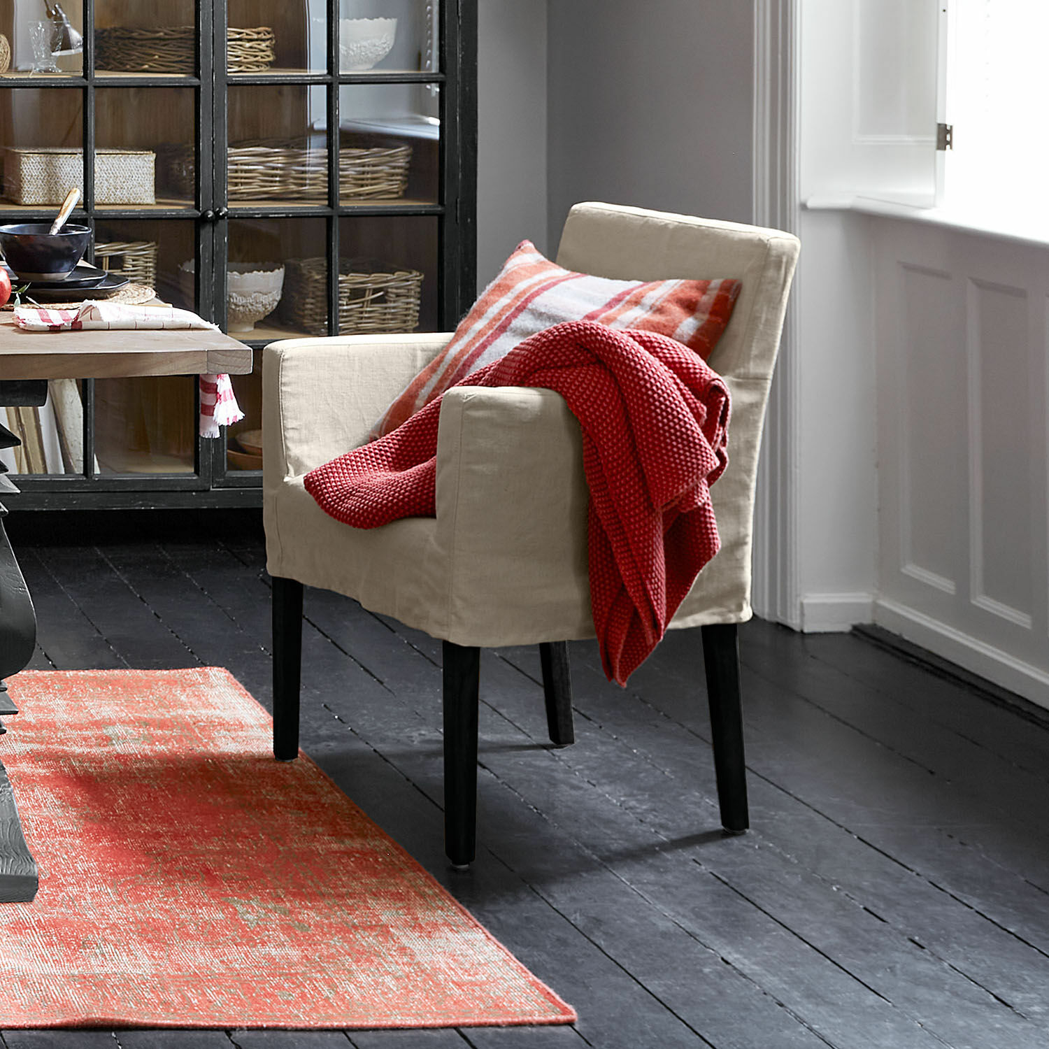 Sedia Longhill Poltrona beige in tessuto con gambe nere, coperta rossa e cuscino a quadri, tappeto rosso