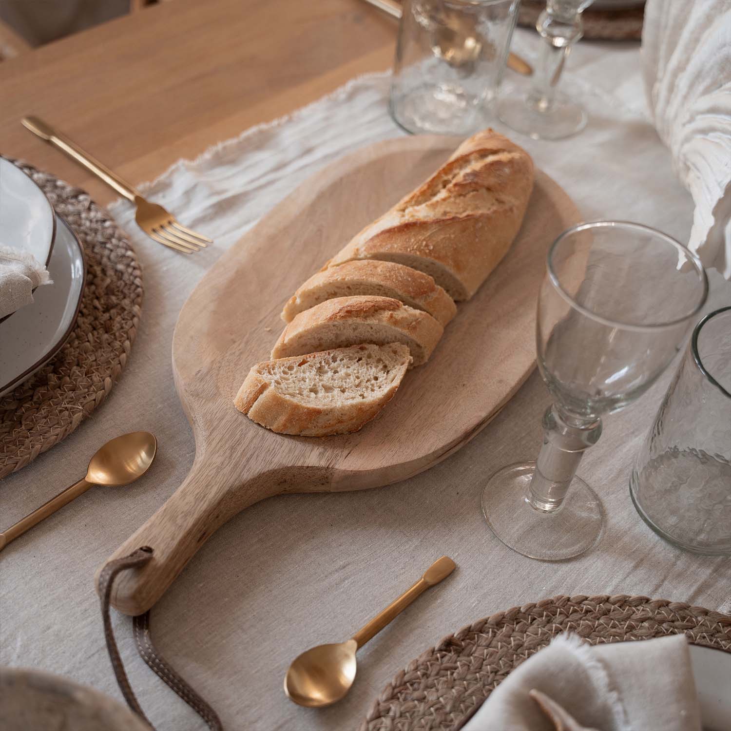 Tagliere in legno con baguette affettata su tavola con tovaglia naturale e posate dorate