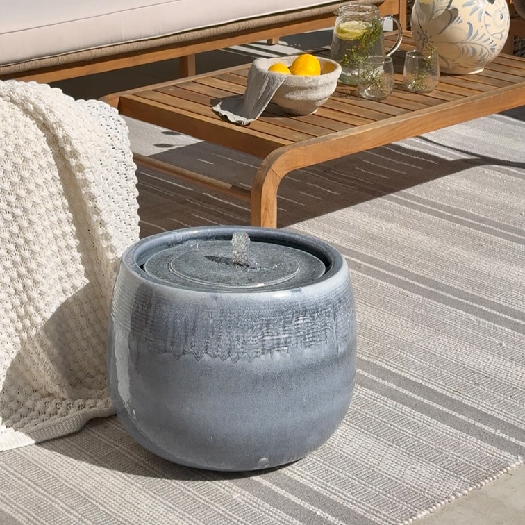 Fontana da tavolo in ceramica grigia su tappeto chiaro accanto a tavolo in legno e coperta bianca