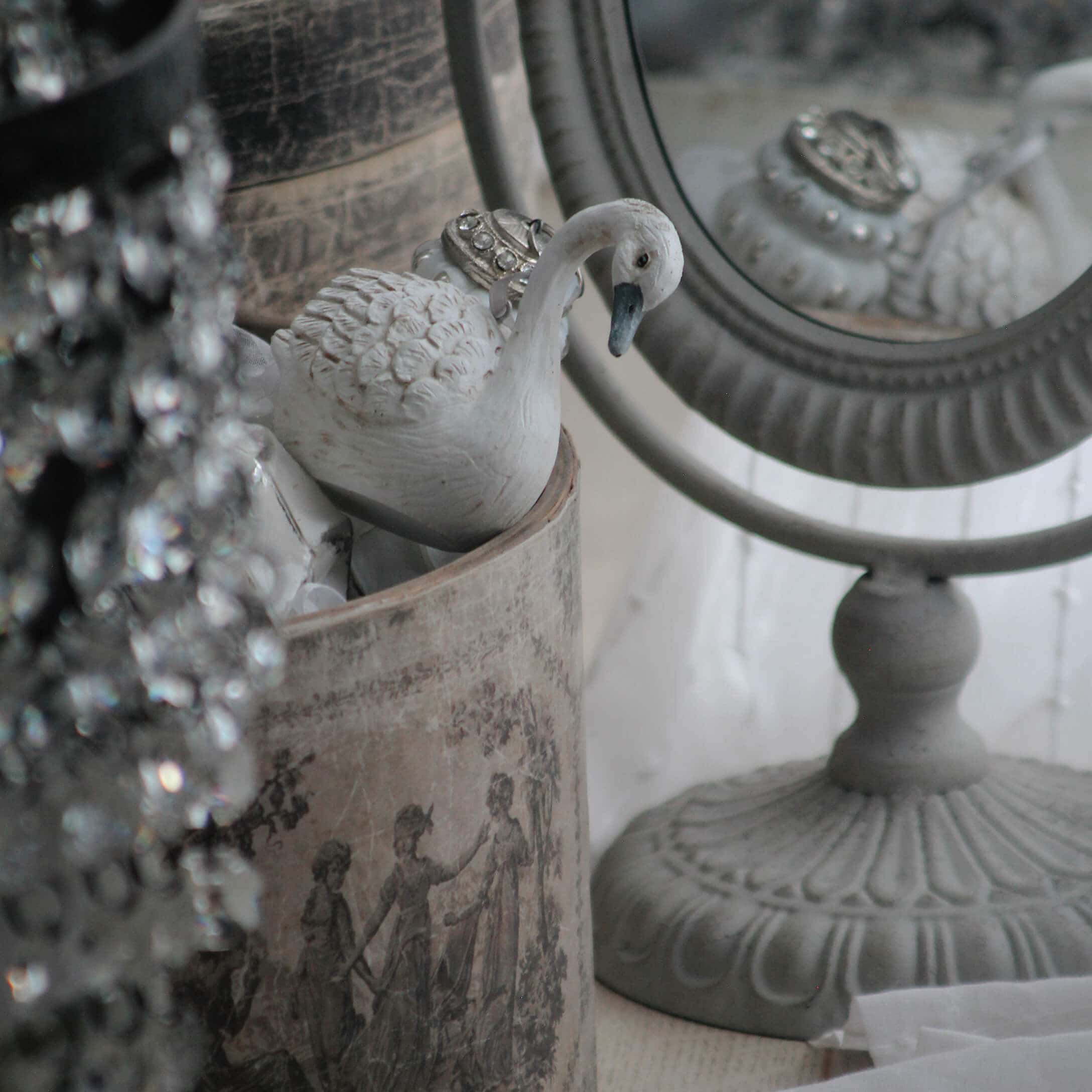 Figura di cigno bianca in ceramica accanto a uno specchio decorativo e un contenitore vintage con motivo classico