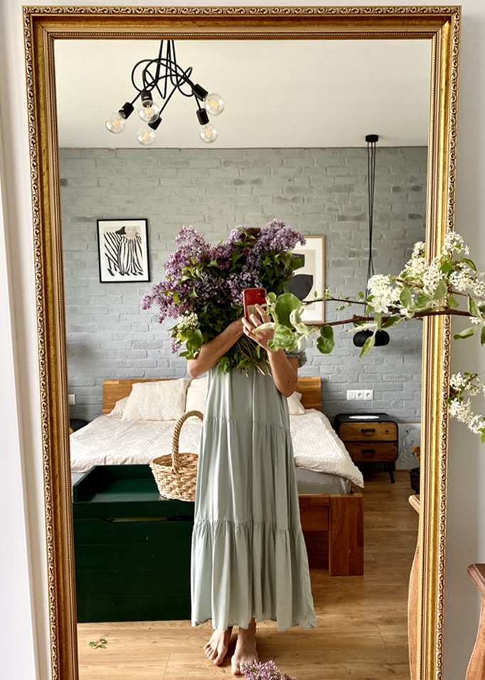 Donna in abito verde lungo che tiene un mazzo di fiori viola davanti a uno specchio in camera da letto con letto in legno