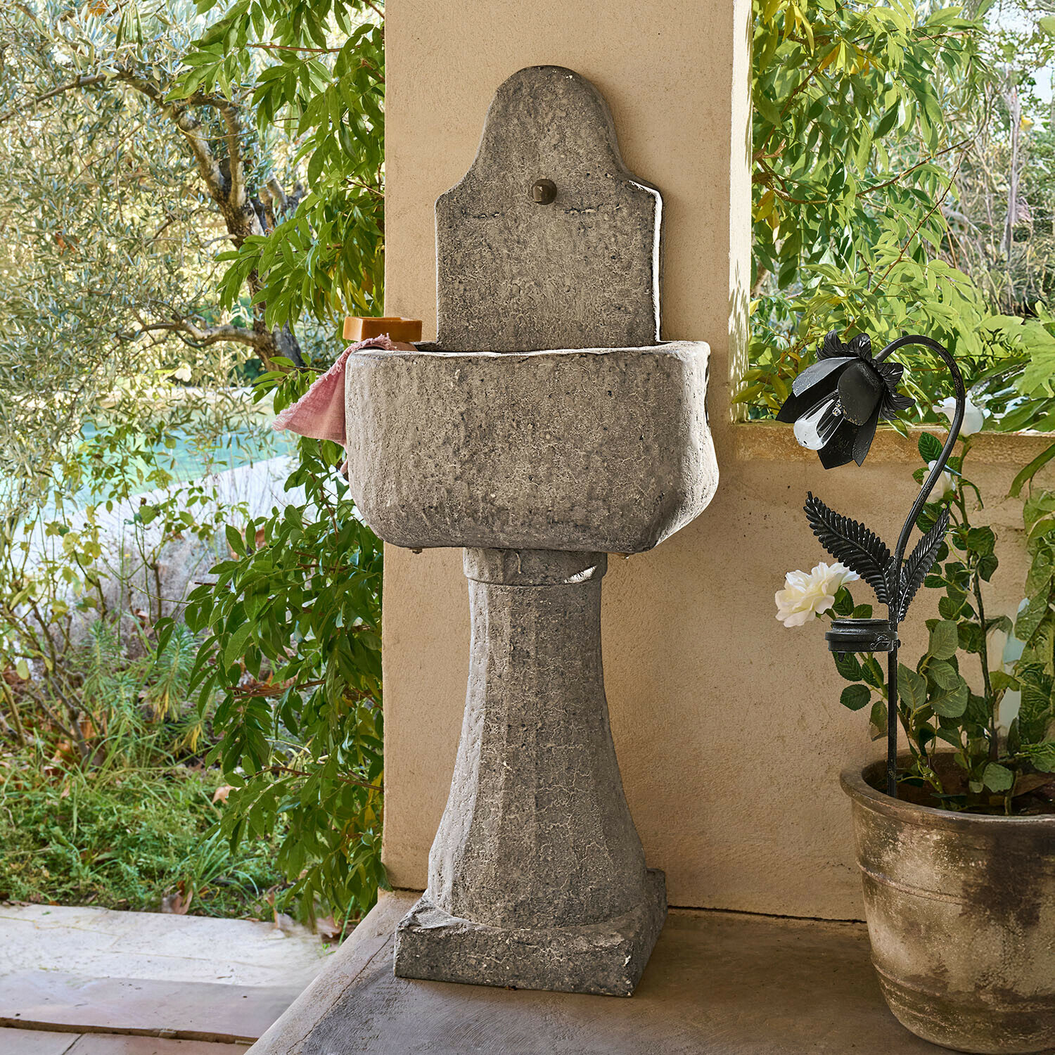 Fontana Lurevino Lavabo da giardino in pietra grigia su colonna con finitura naturale e piante verdi sullo sfondo