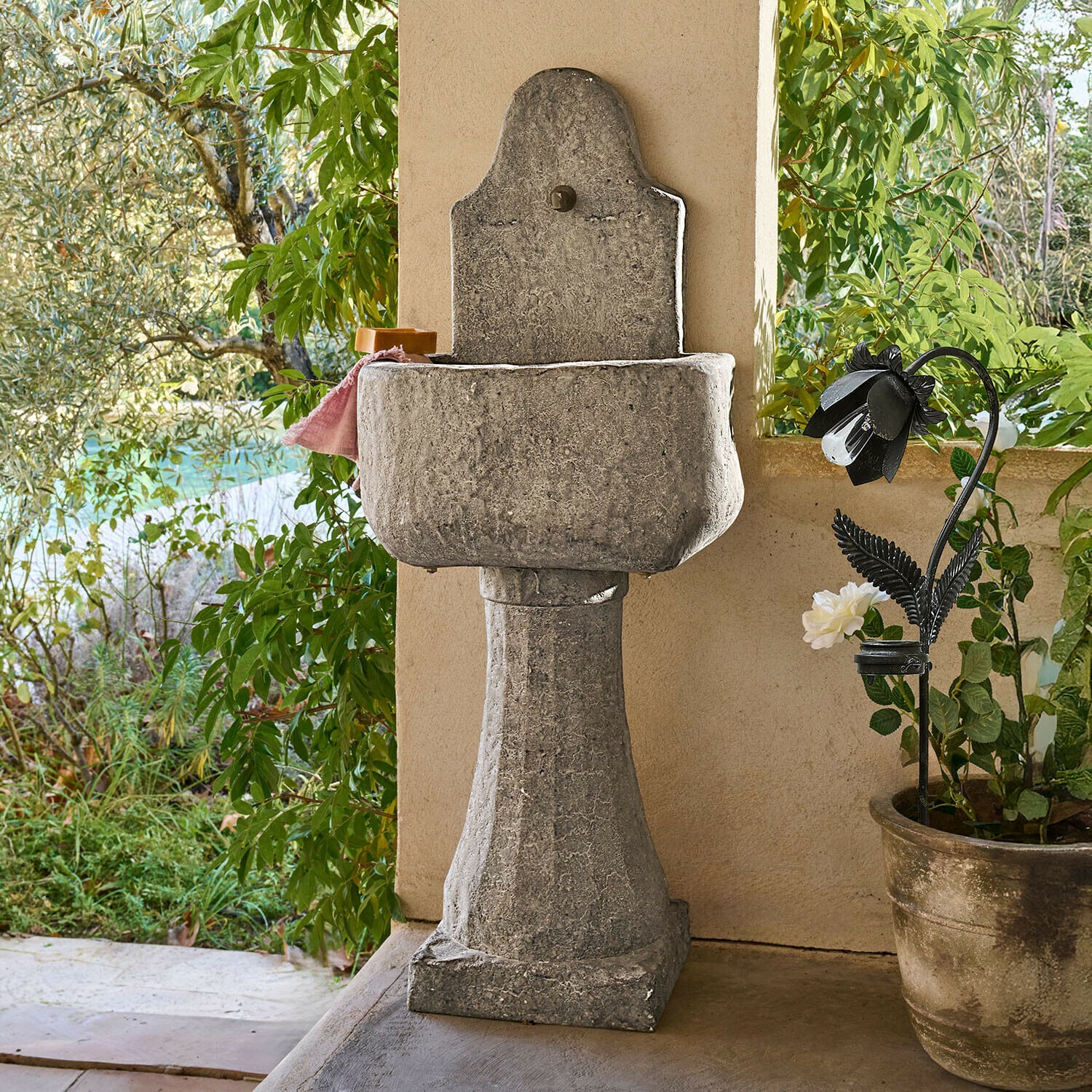 Fontana Lurevino Lavabo da giardino in pietra grigia su colonna con finitura naturale e piante verdi sullo sfondo