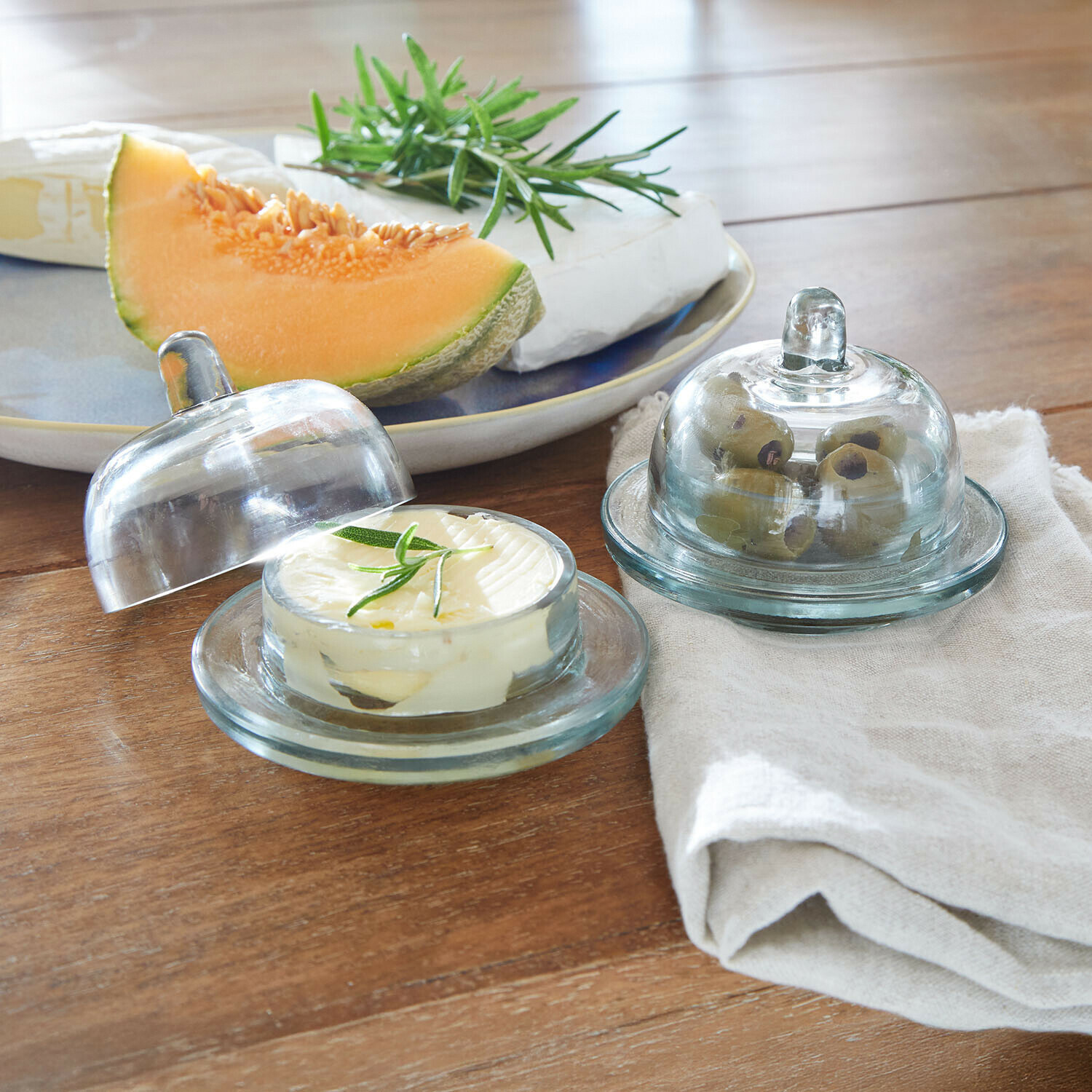 Coppia di portaburro Doraville Contenitori in vetro per burro e olive su tavolo in legno con melone e formaggio sullo sfondo