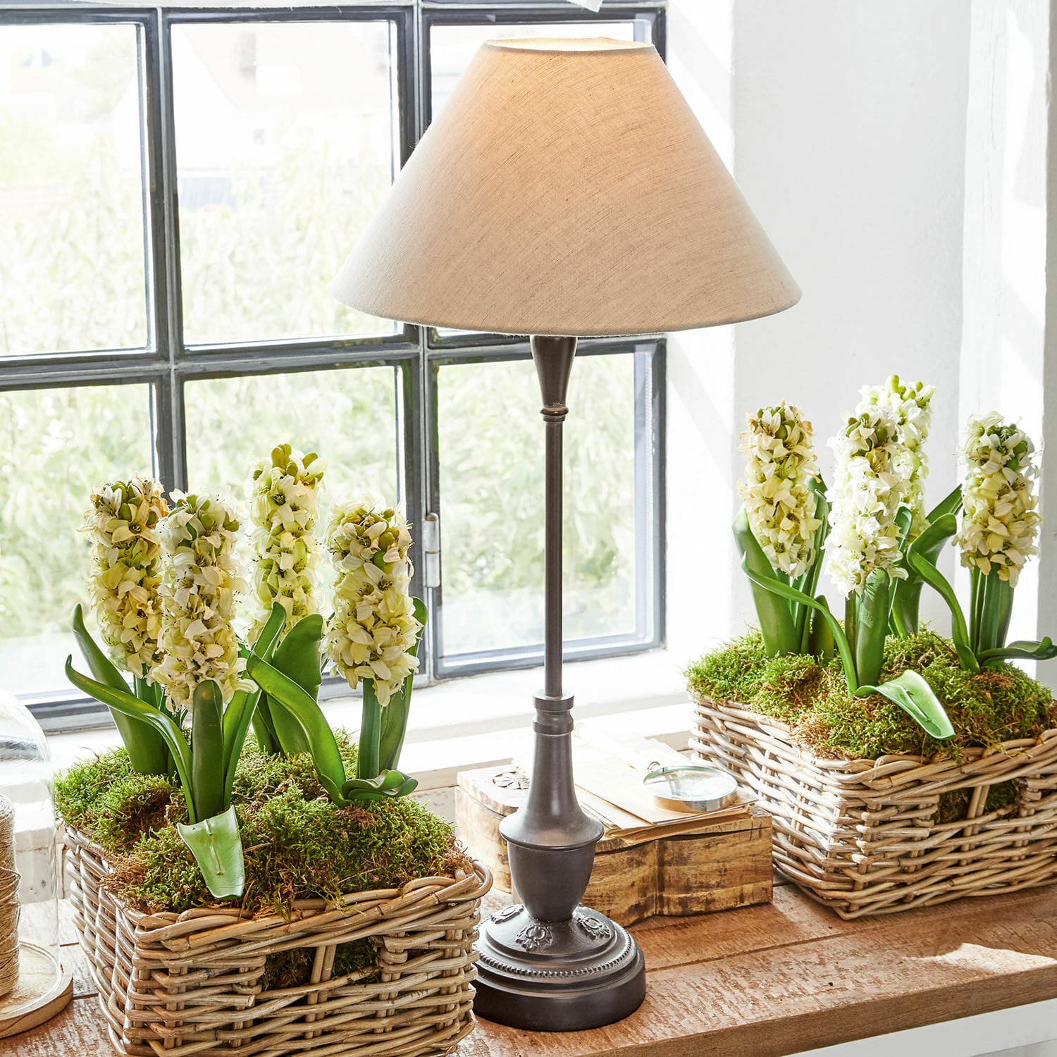 Lampada da tavolo Barnwell Lampada da tavolo con base in metallo e paralume beige accanto a cesti di vimini con fiori bianchi e muschio