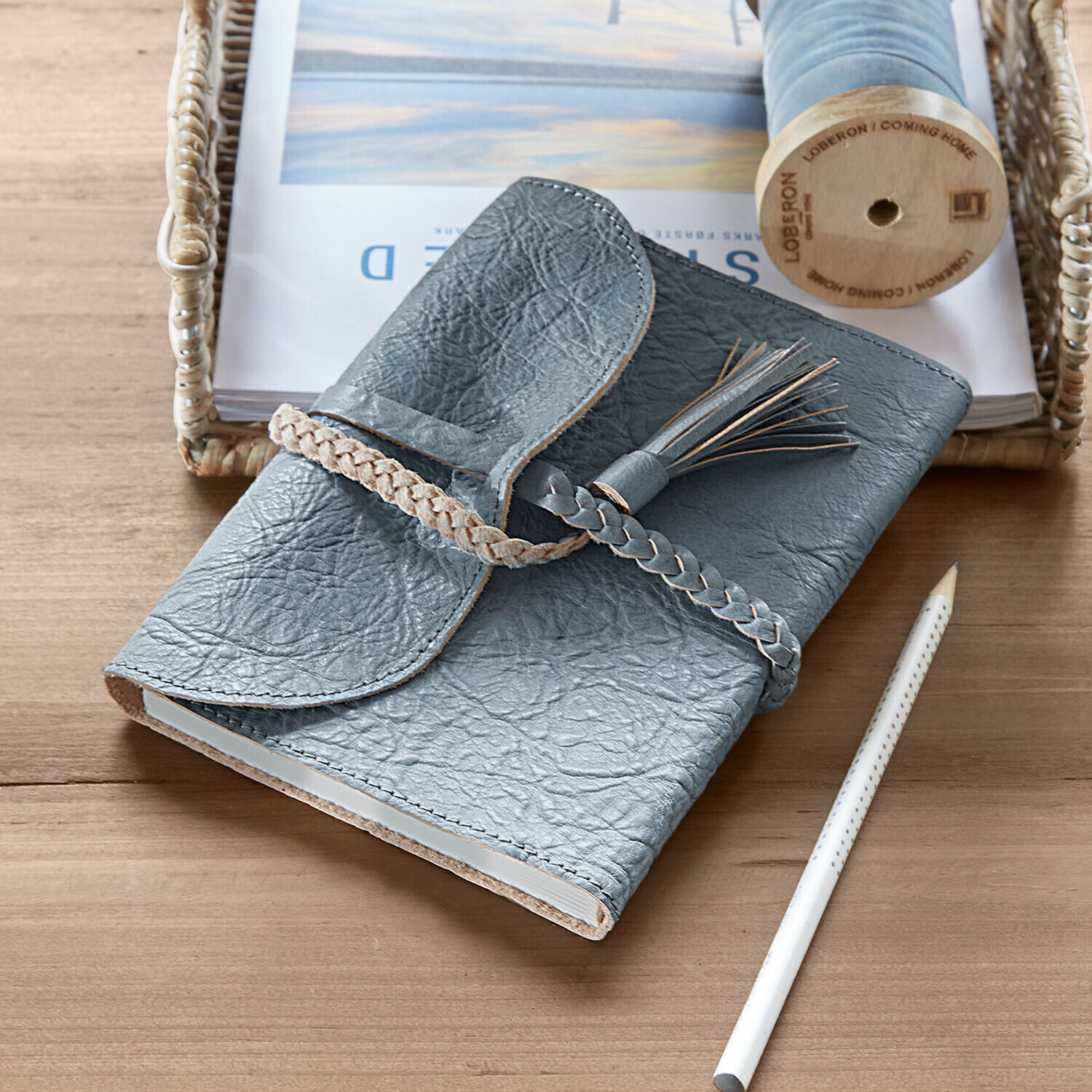 Taccuino in pelle grigia con chiusura intrecciata e nappina su tavolo di legno con matita bianca