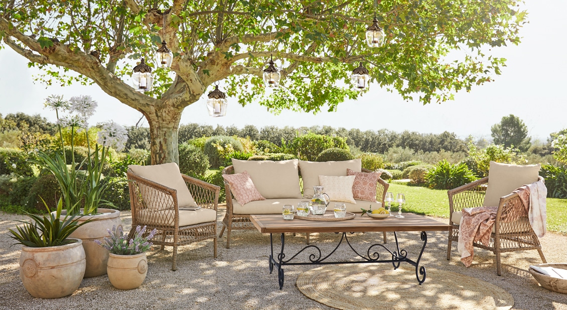Set di mobili da giardino con cuscini beige e tavolo in legno sotto un albero in terrazza