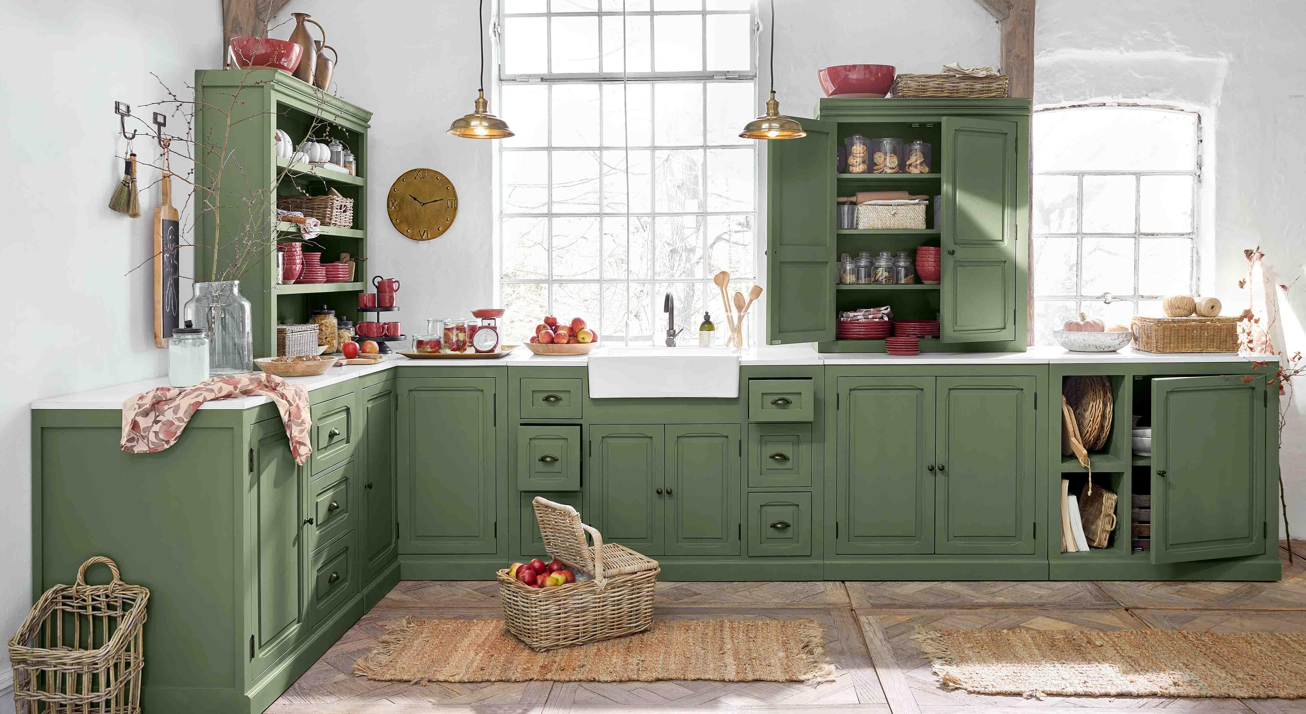 Cucina con mobili in legno verde e piano bianco, accessori rustici e cesta di mele