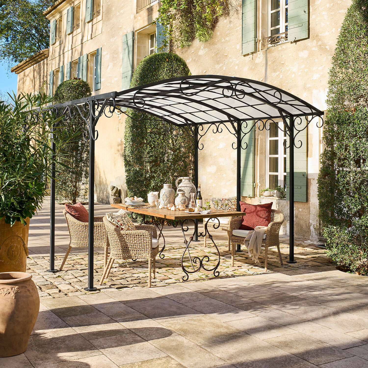 Gazebo in metallo nero con decorazioni a volute sopra tavolo in legno e sedie in vimini