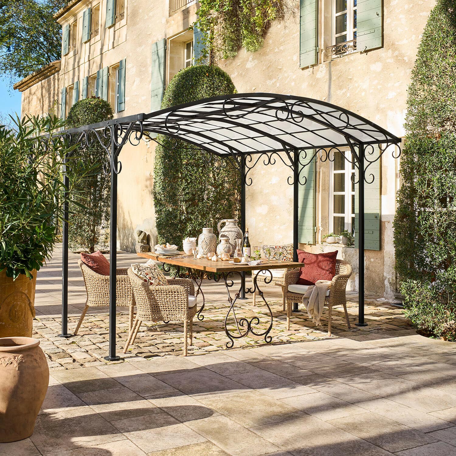 Pergola Delbridge Gazebo in metallo nero con decorazioni a volute sopra tavolo in legno e sedie in vimini