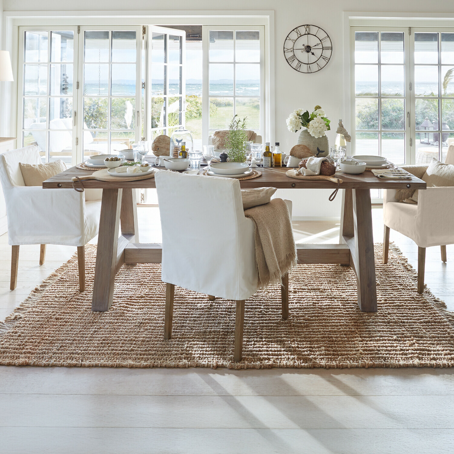 Tavolo Berrien Sala da pranzo con tavolo in legno e sedie bianche su tappeto naturale con vista su giardino e mare