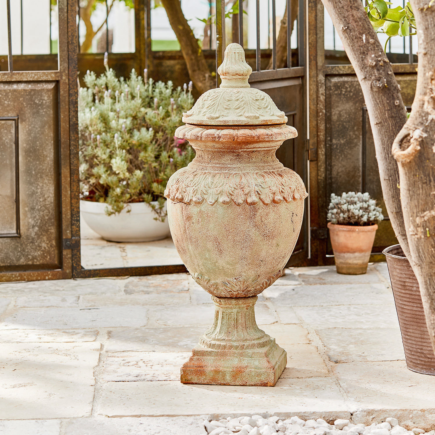 Anfora decorativa Persephone Vaso decorativo da giardino in terracotta colore naturale su pavimento in pietra con piante sullo sfondo