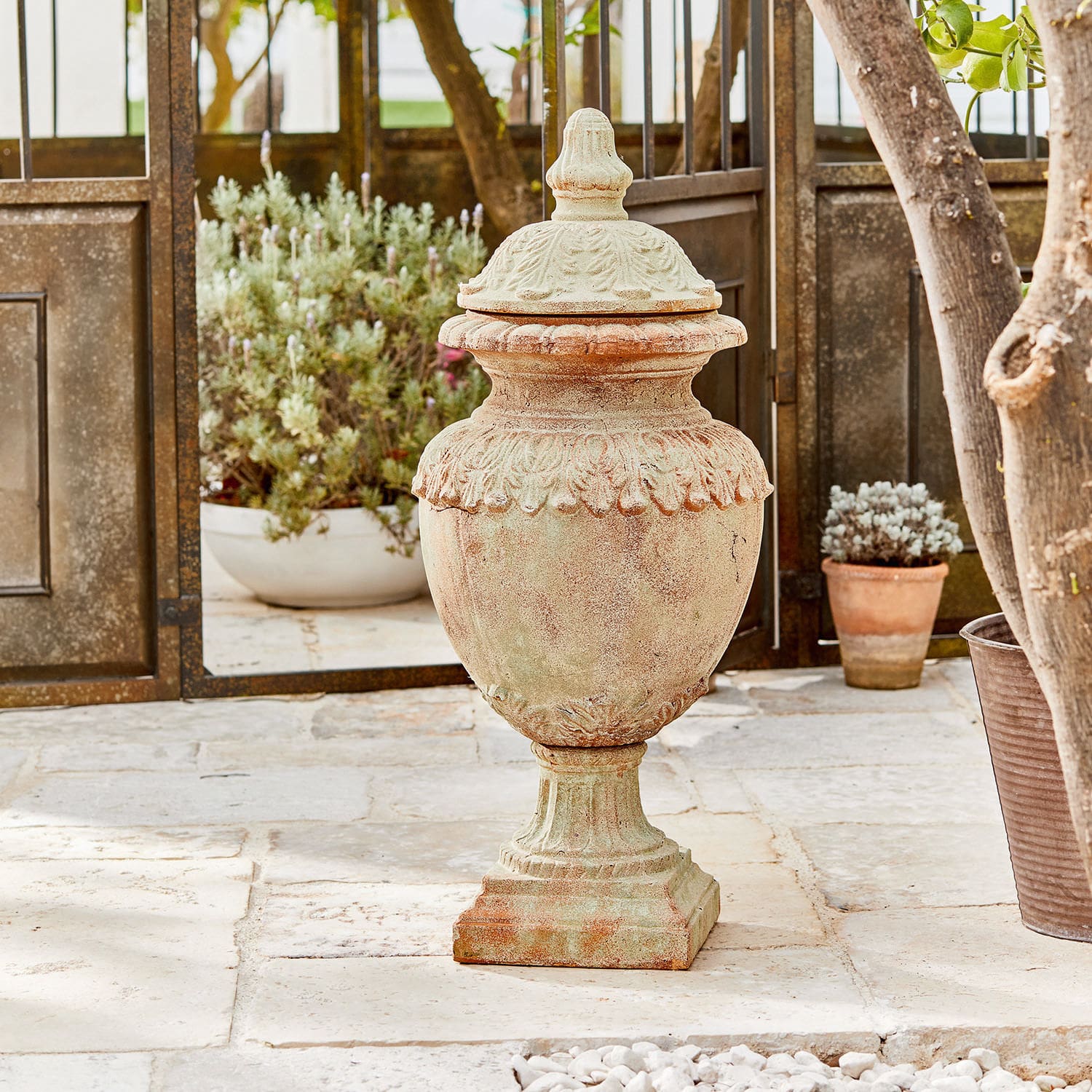 Anfora decorativa Persephone Vaso decorativo da giardino in terracotta colore naturale su pavimento in pietra con piante sullo sfondo