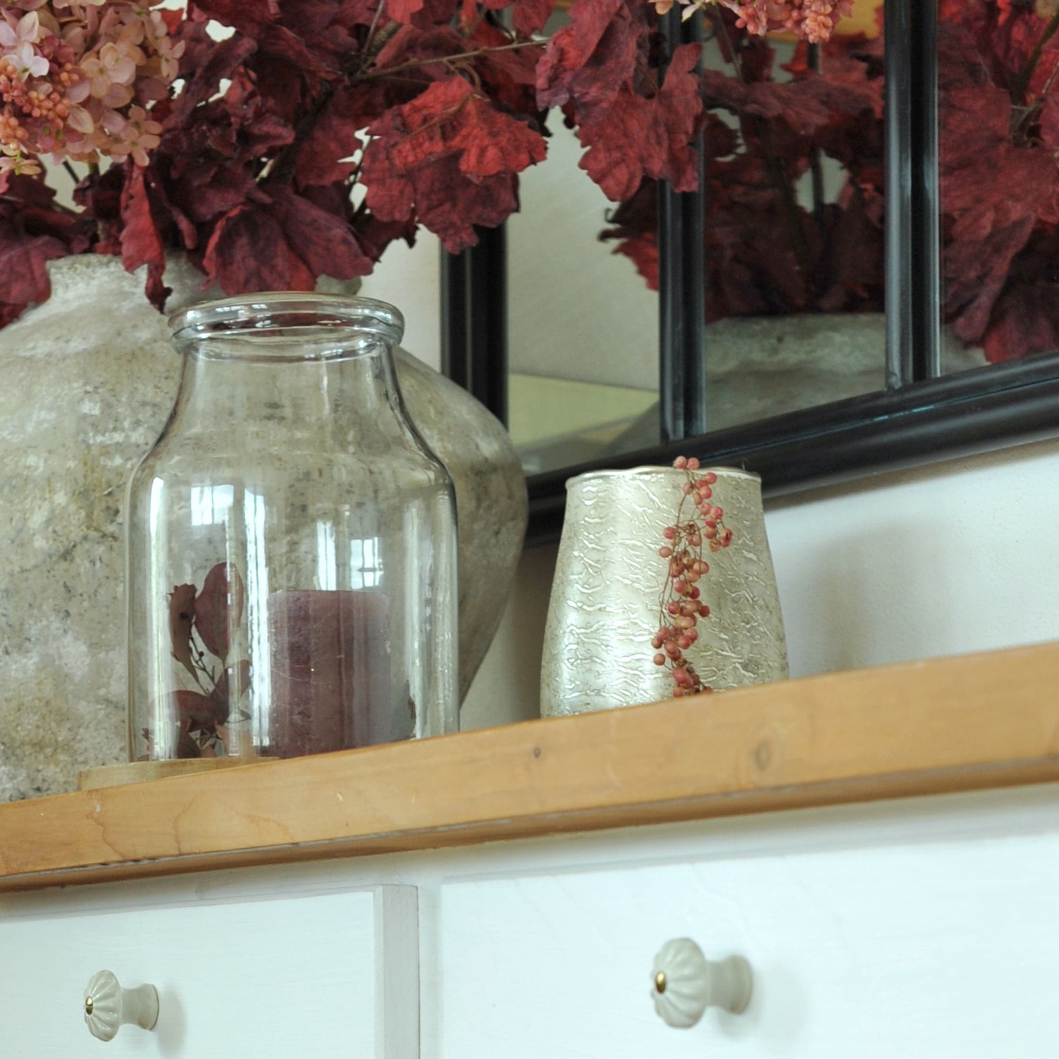 Vaso in vetro trasparente con candela bordeaux su mensola in legno con decorazione di foglie rosse