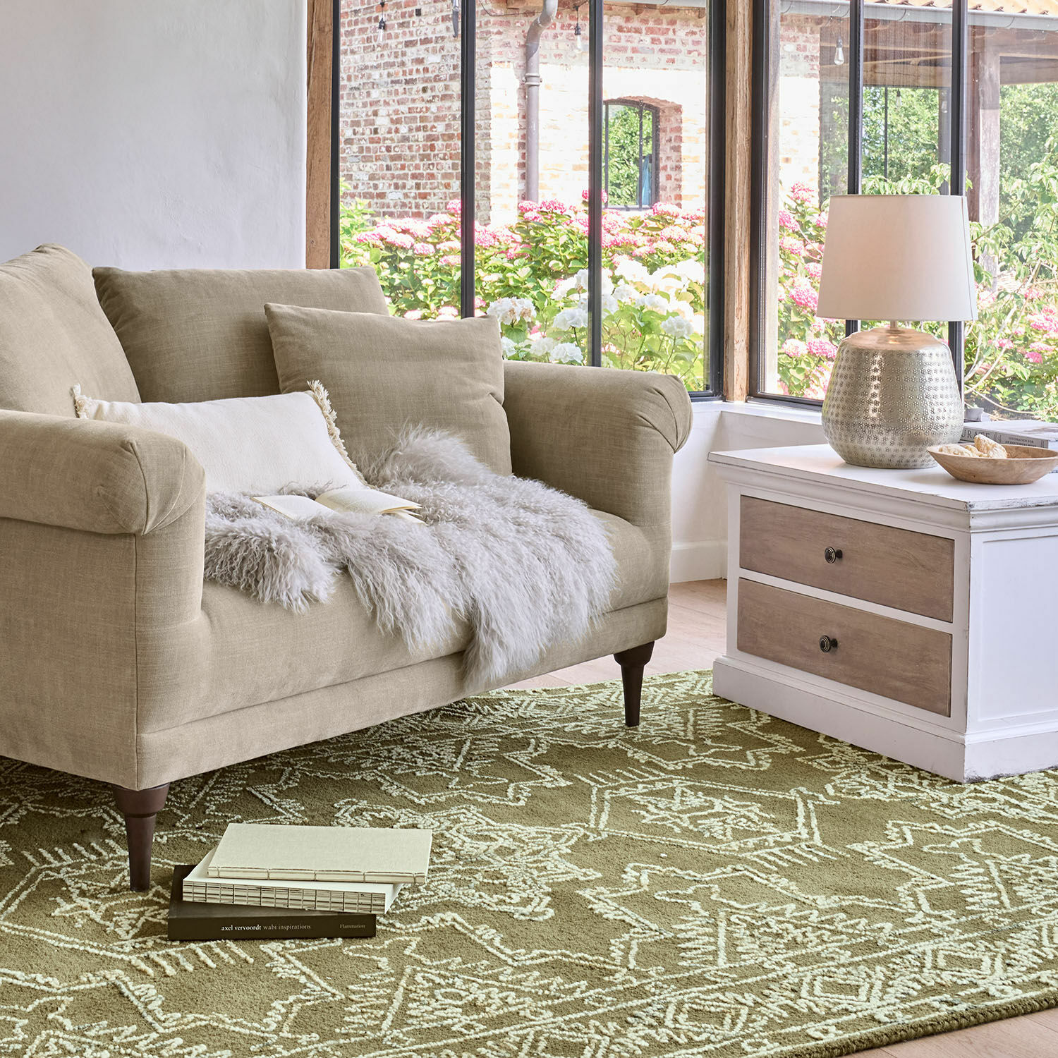 Tappeto Qazoura Divano beige in tessuto con cuscini e coperta di pelliccia su tappeto verde con motivo geometrico