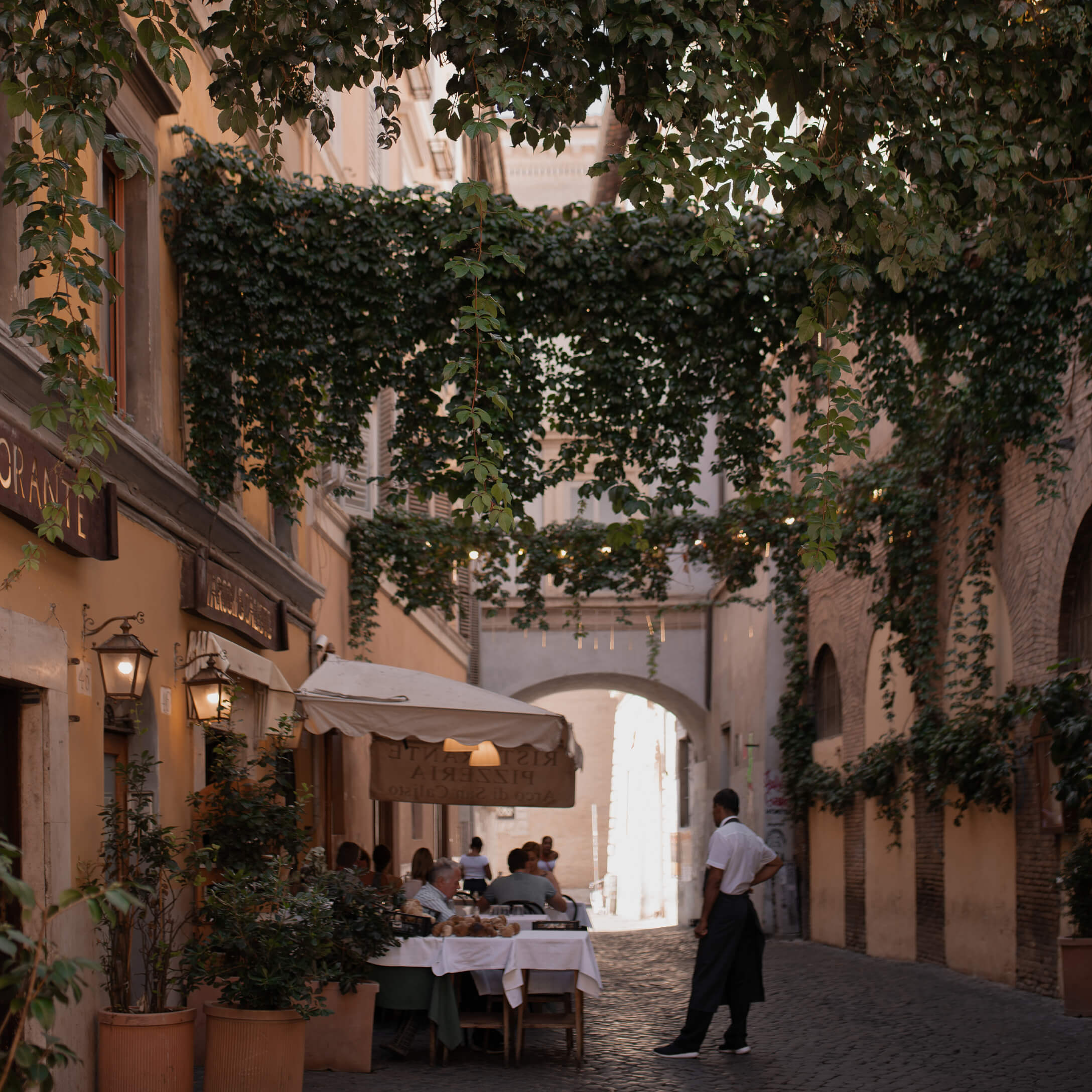 Stradina lastricata con ristorante e terrazza sotto piante rampicanti verdi, atmosfera accogliente
