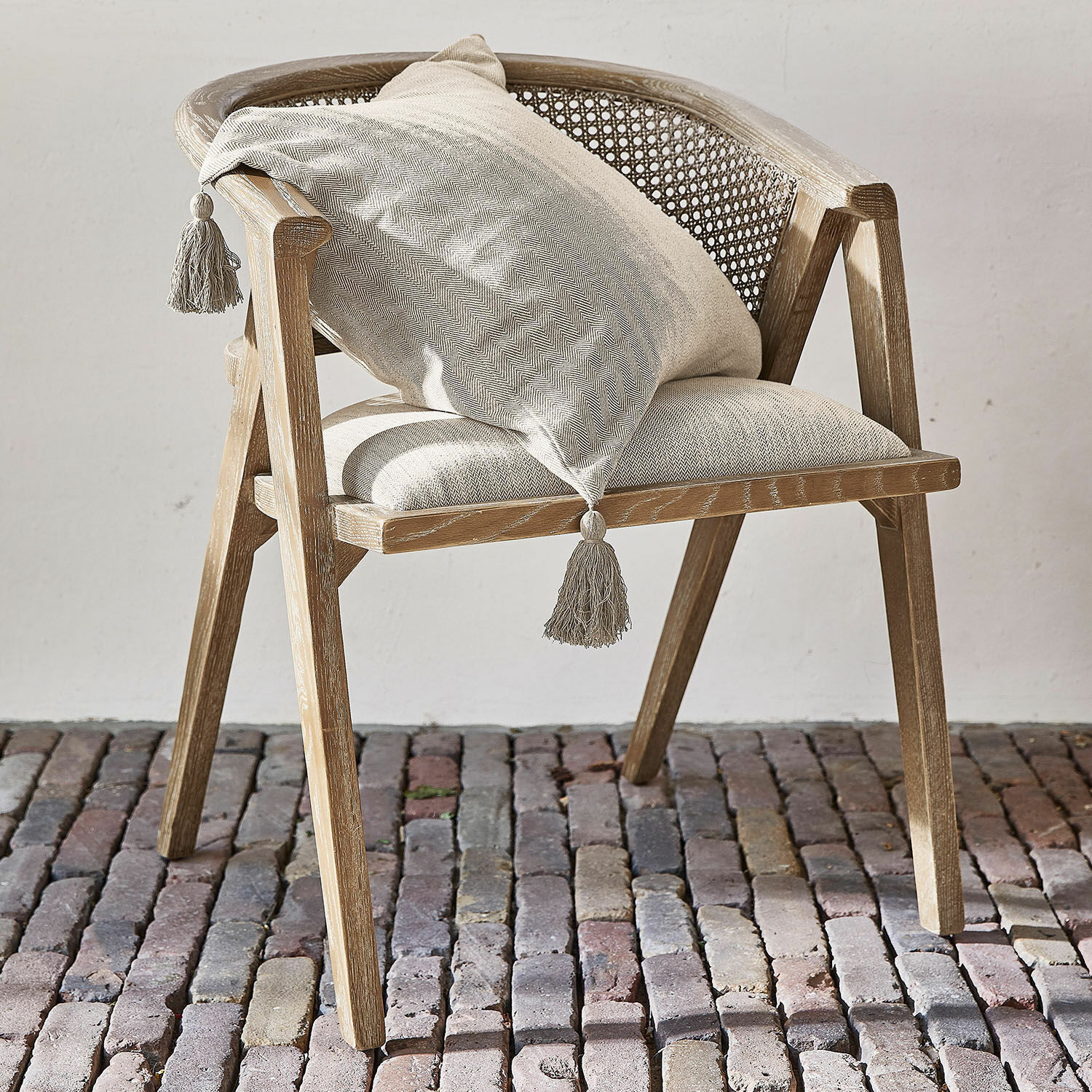 Sedia Collaroy Sedia in legno chiaro con schienale in vimini e cuscino beige con nappine su pavimento in pietra