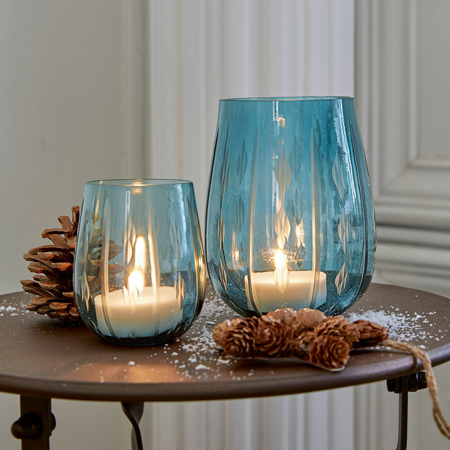 Due lanterne in vetro blu con motivo, candele bianche accese e decorazione di pigne sul tavolo