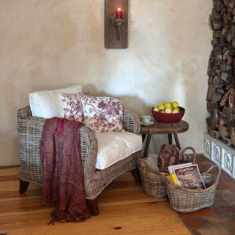 Poltrona in vimini con cuscino bianco e coperta bordeaux accanto a tavolino in legno con frutta e cesti