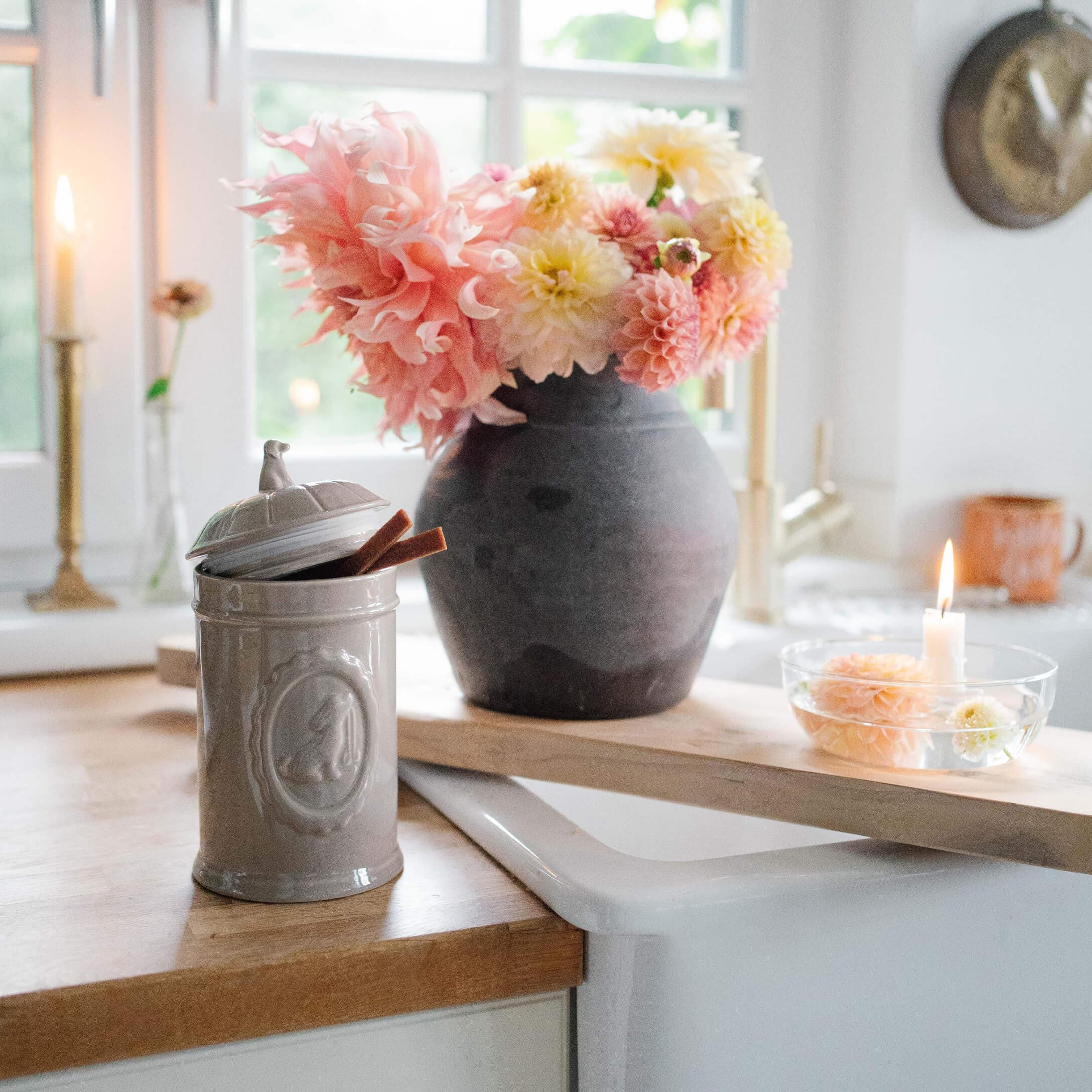 Barattolo in ceramica grigia con coperchio su piano in legno accanto a vaso nero con fiori rosa e gialli