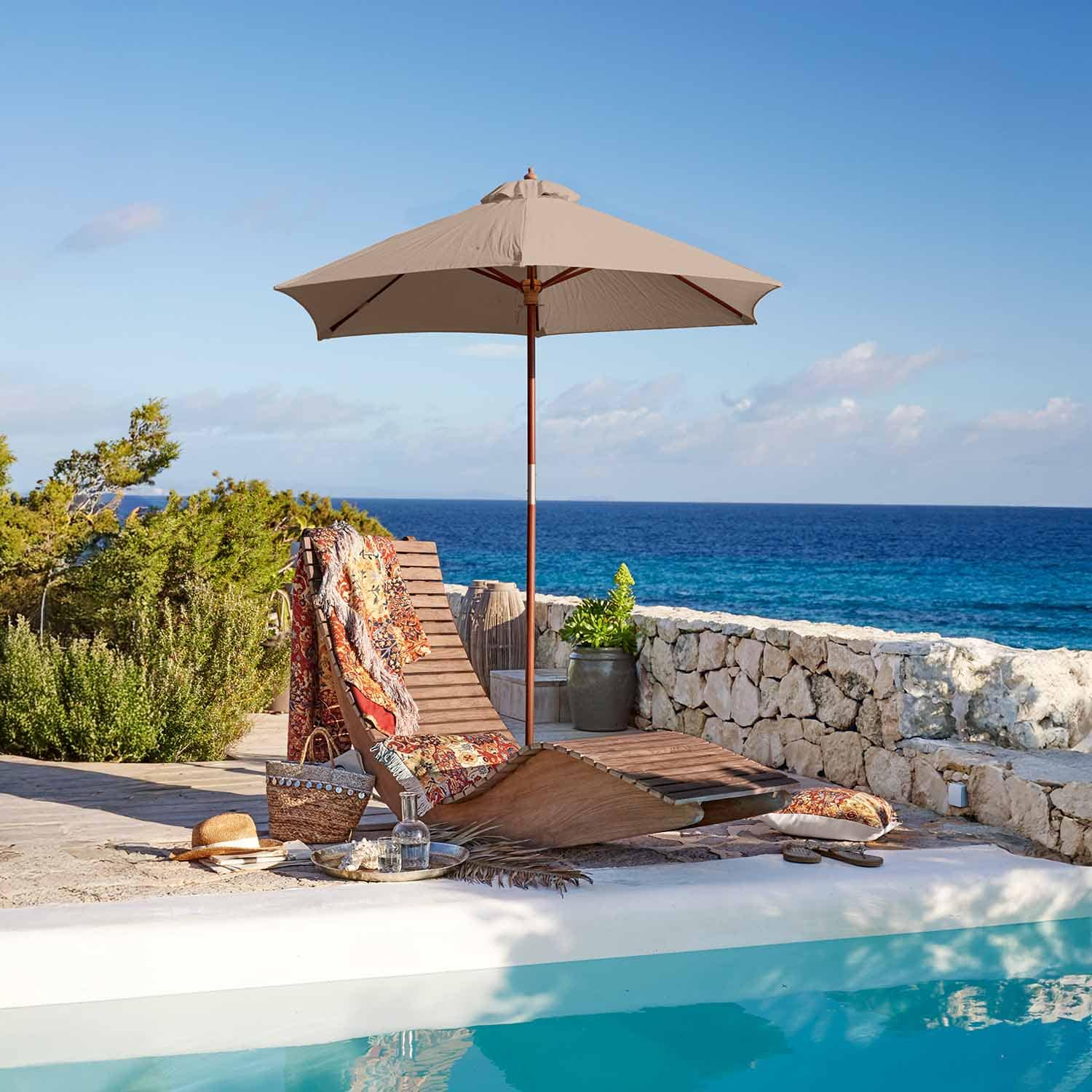 Lettino in legno con ombrellone beige a bordo piscina con vista mare e coperta colorata