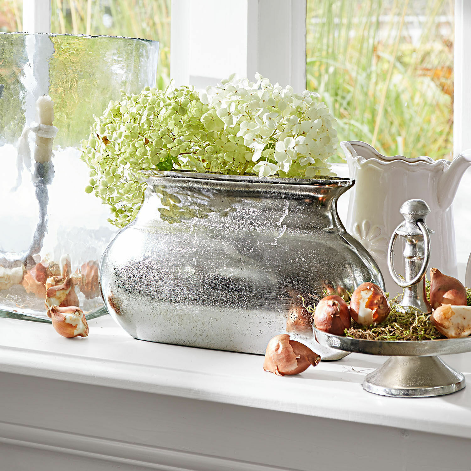 Vaso Melisique Vaso metallico argentato con ortensie verde e bianco su davanzale con bulbi decorativi