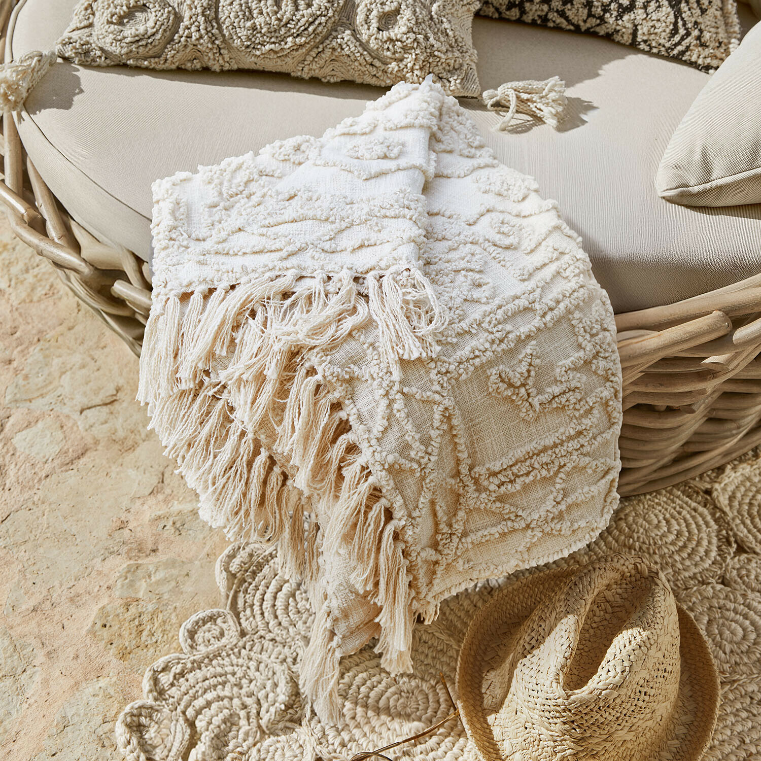 Coperta decorativa beige con frange su poltrona in vimini e cappello di paglia sul pavimento