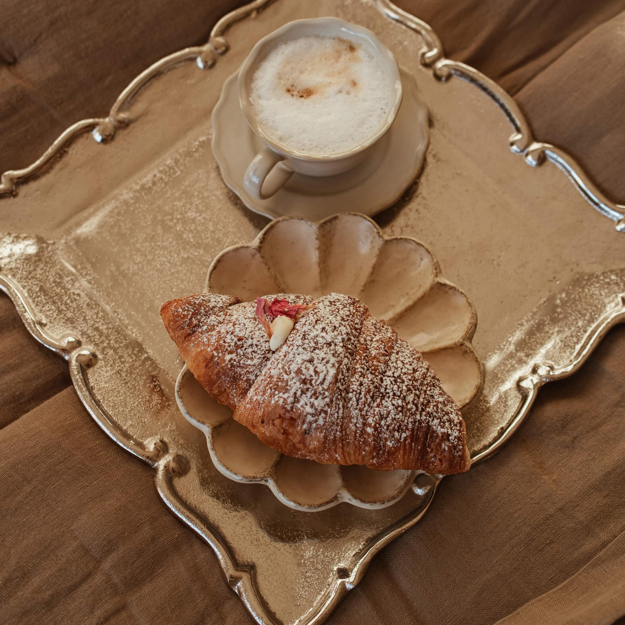 Tazza di cappuccino cremoso e croissant con zucchero a velo su piatto beige e vassoio dorato