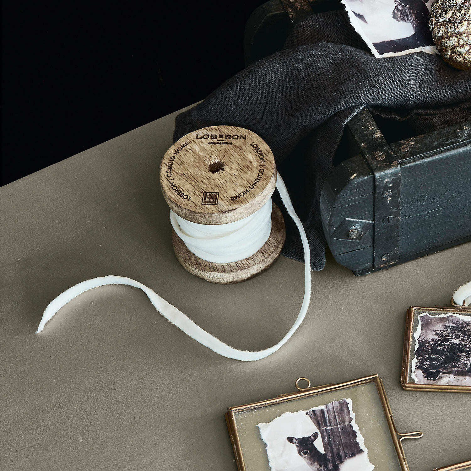 Nastro di velluto Olessa Bobina in legno con tessuto bianco su tavolo accanto a scatola nera e cornici decorative con foto