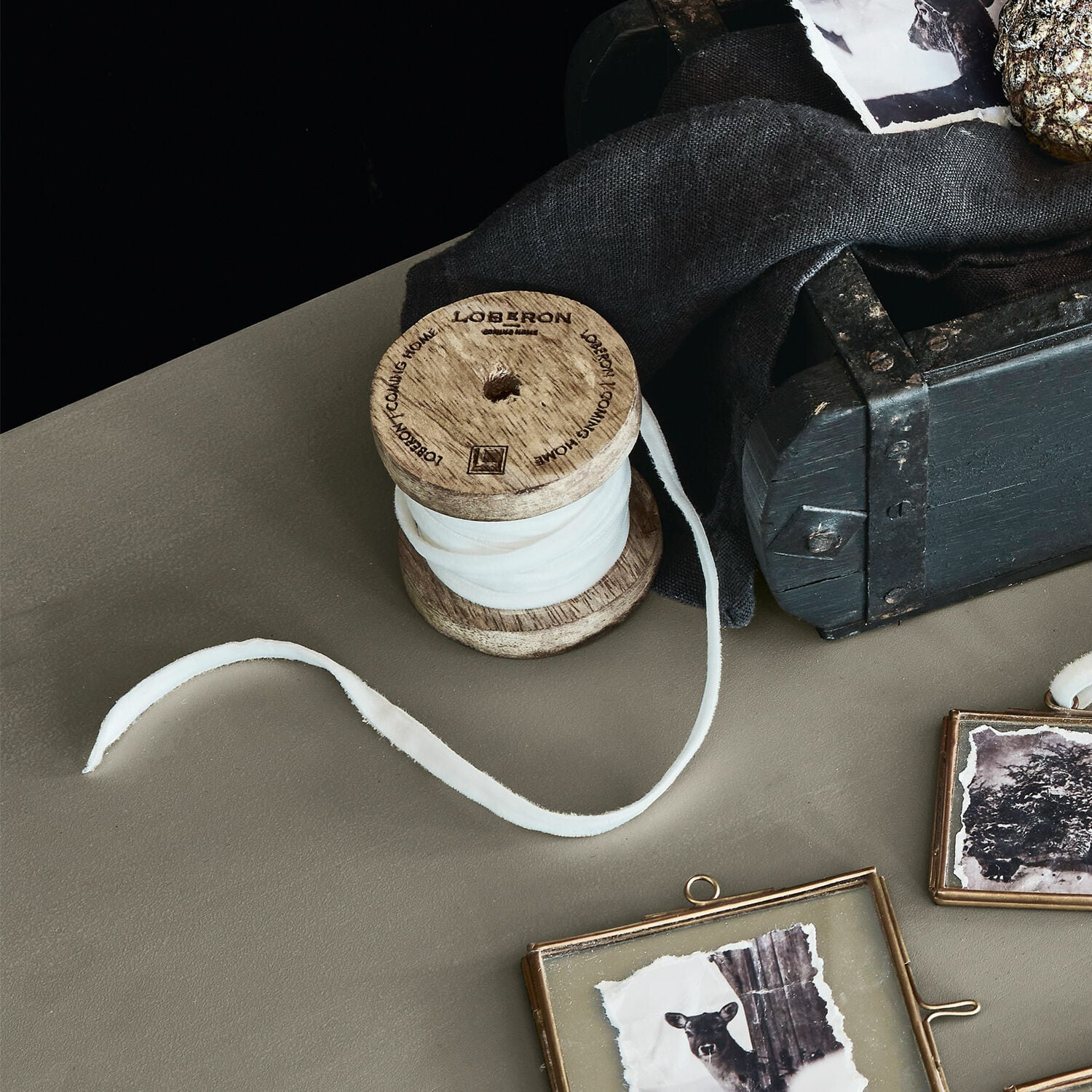 Nastro di velluto Olessa Bobina in legno con tessuto bianco su tavolo accanto a scatola nera e cornici decorative con foto