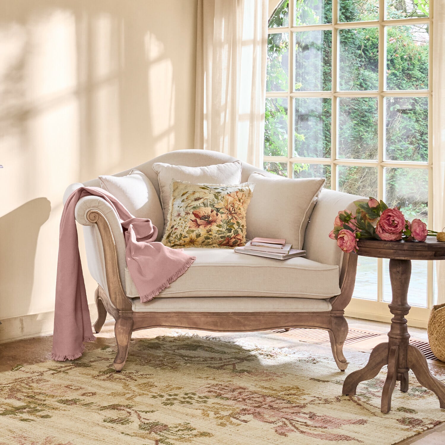 Divano beige con struttura in legno, cuscino floreale e coperta rosa in soggiorno luminoso con finestra