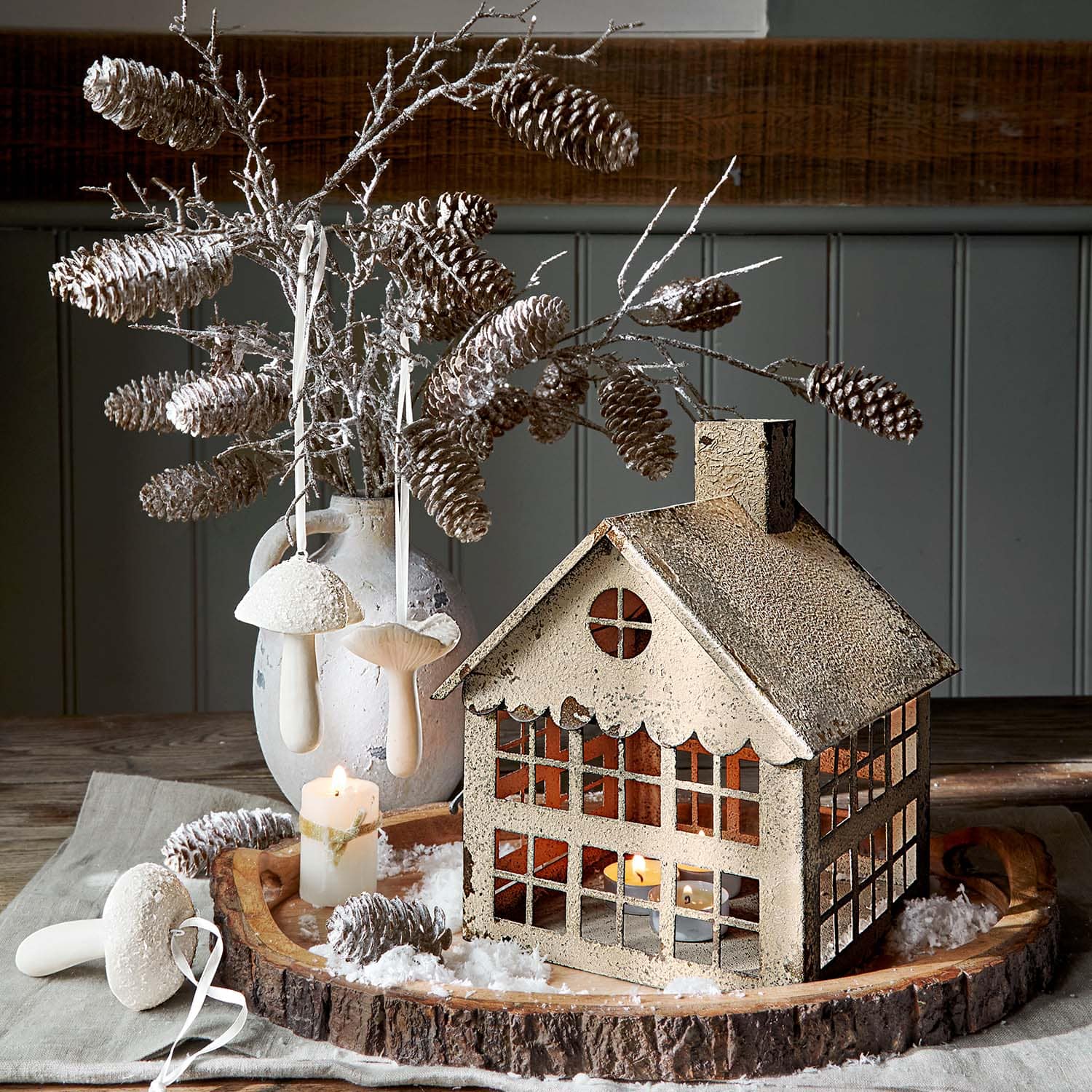 Lanterna decorativa in metallo beige a forma di casa con rami di pigne e candela su vassoio di legno