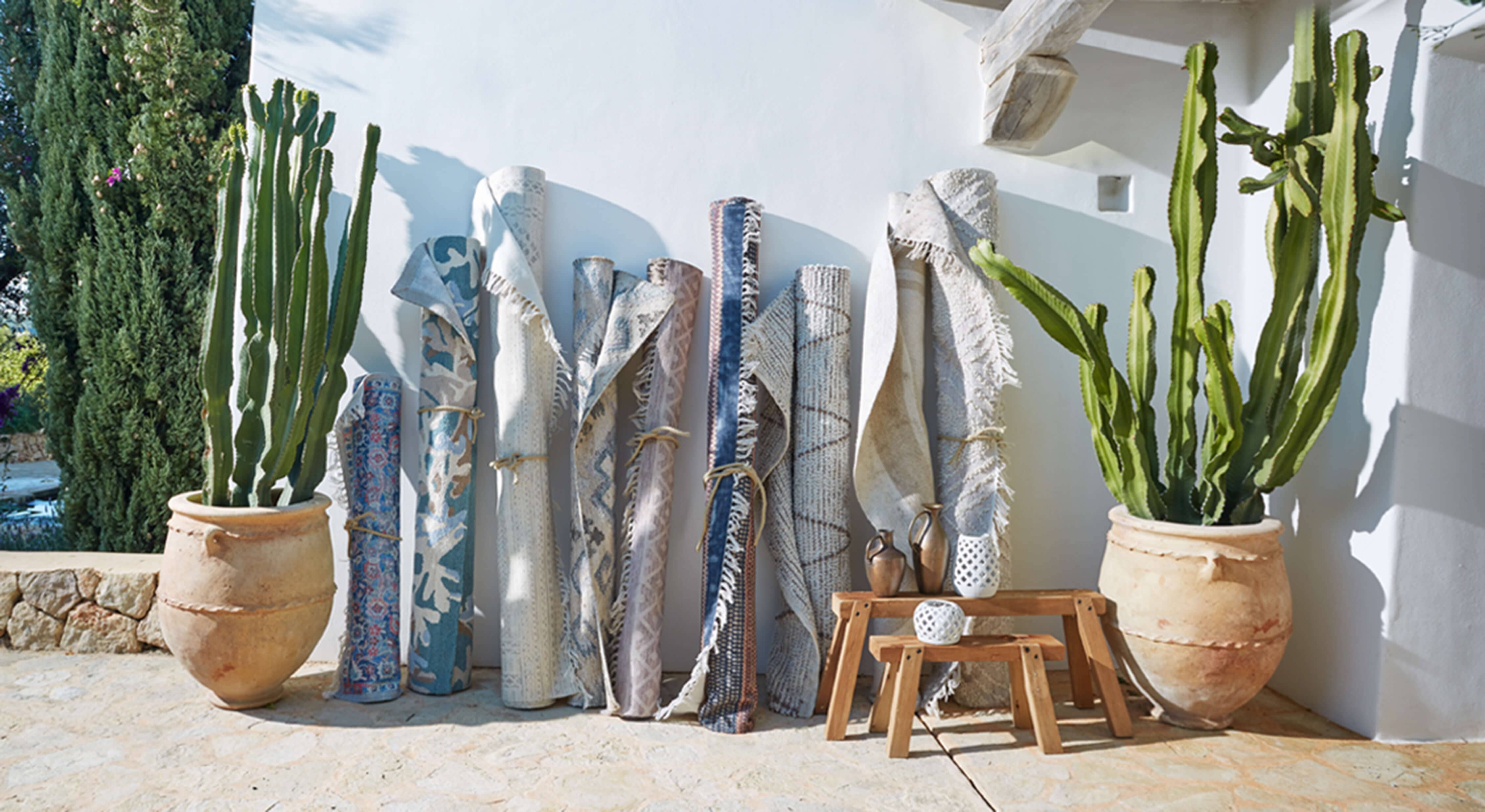 Set di tappeti arrotolati in tonalità blu e beige appoggiati a muro bianco con grandi vasi e cactus