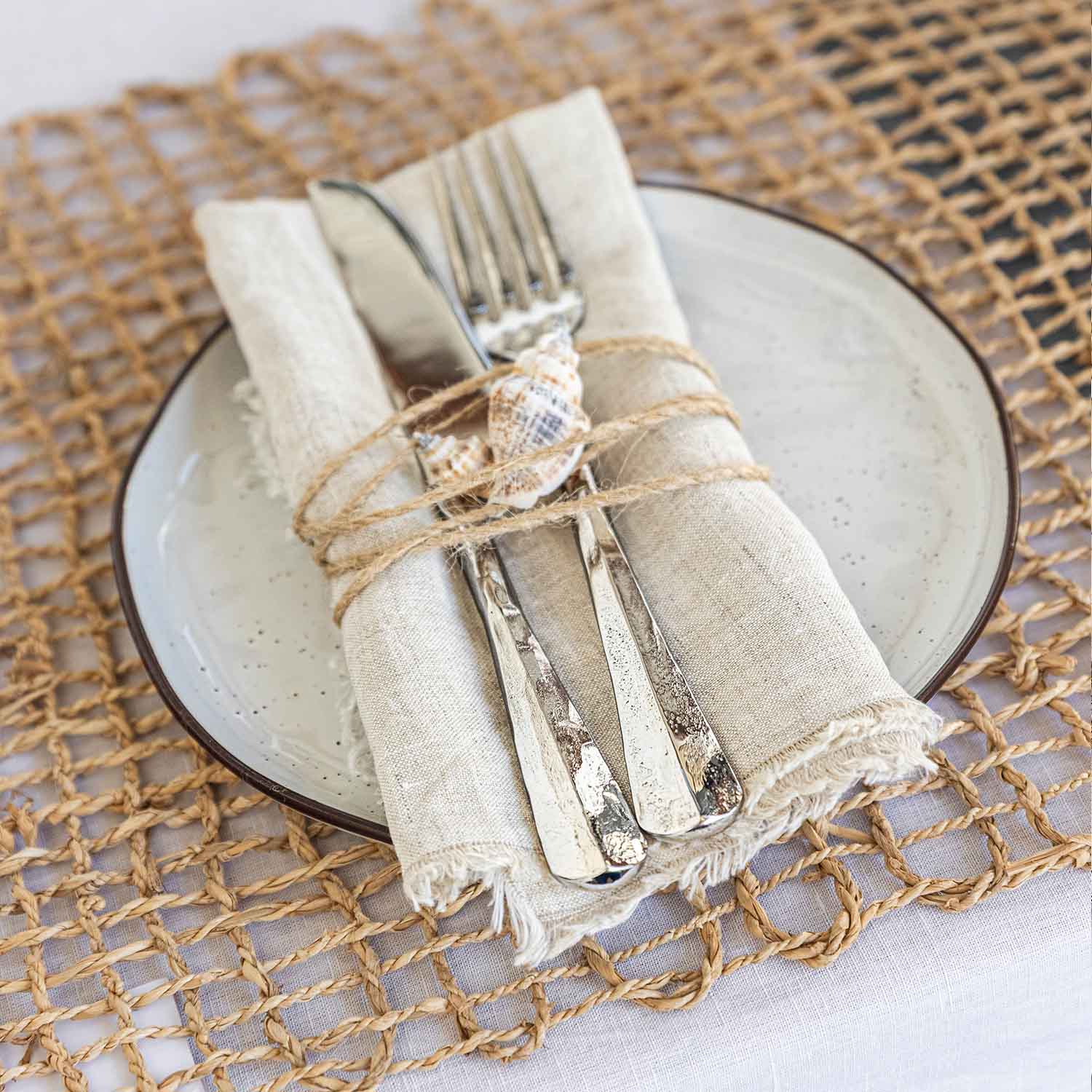 Posate in acciaio inox su tovagliolo di lino con conchiglia, piatto in ceramica e sottopiatto intrecciato