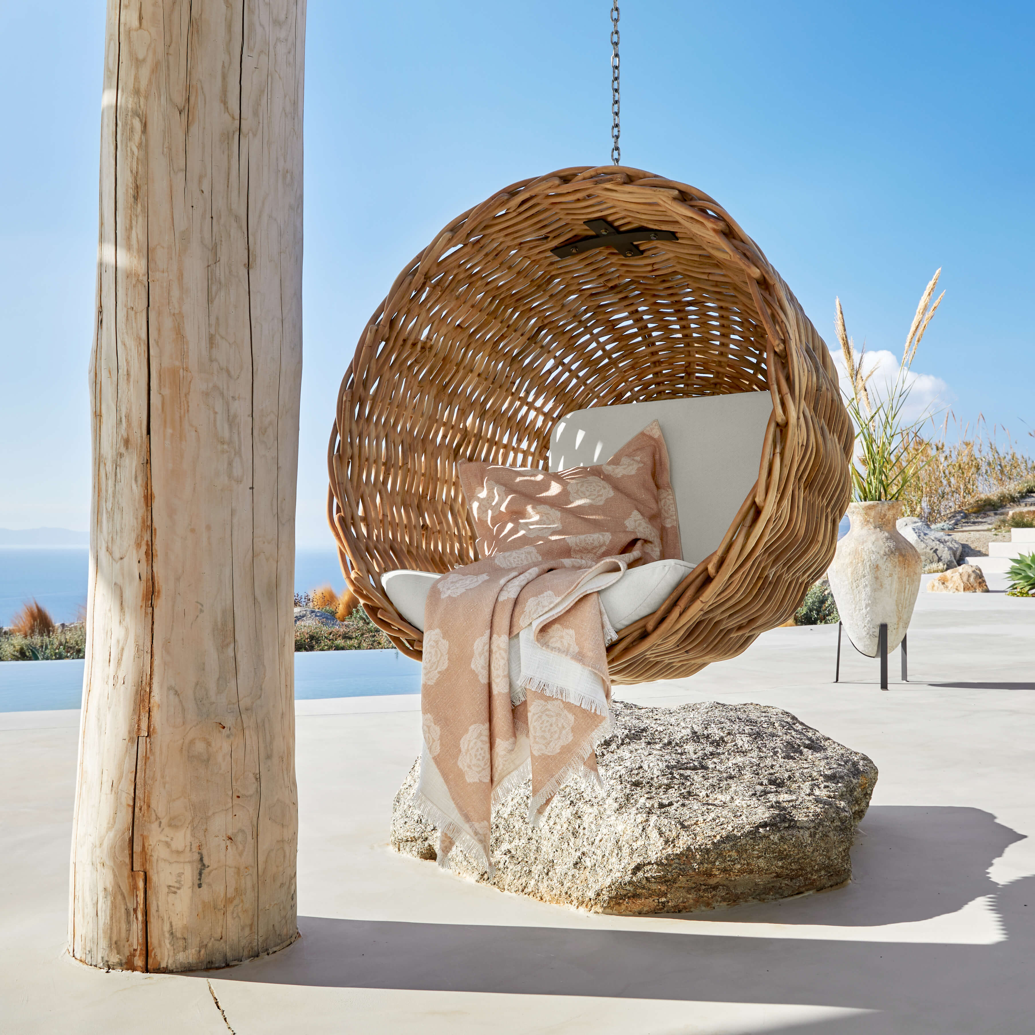 Sedia sospesa in rattan naturale con cuscino chiaro e coperta beige su terrazza con vista mare