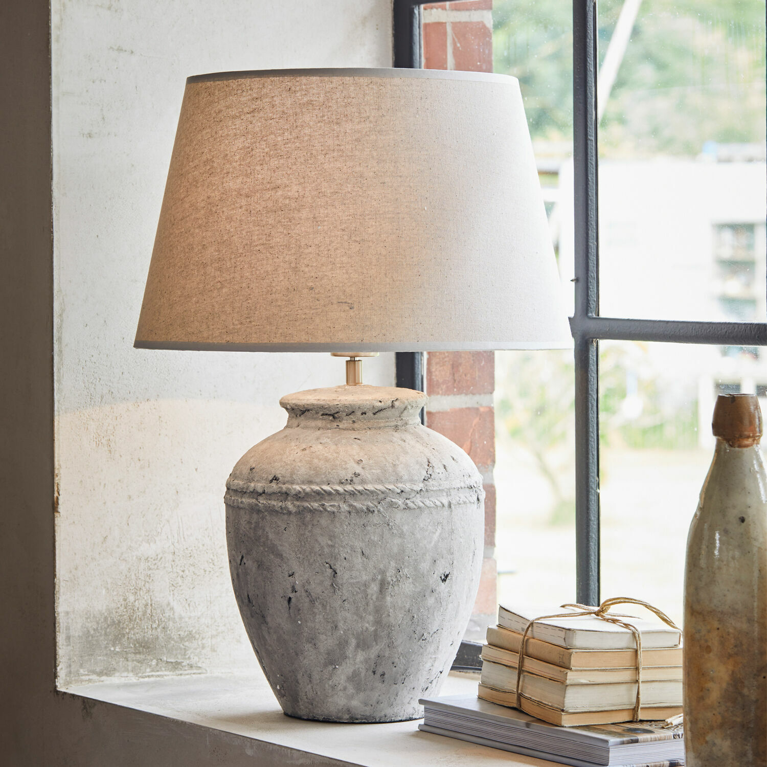 Lampada da tavolo Mosnay Lampada da tavolo in ceramica grigia con paralume beige sul davanzale accanto a libri