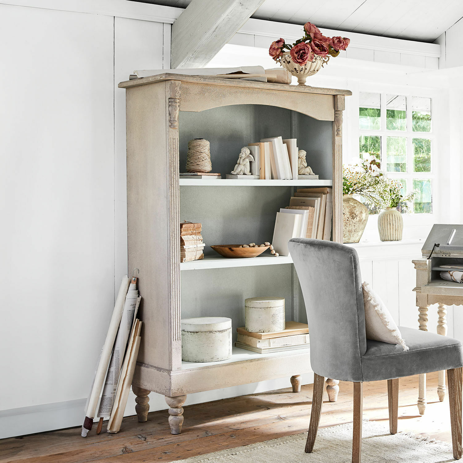 Scaffale Arterton Libreria vintage in legno beige con libri decorativi e sedia grigia in stanza luminosa