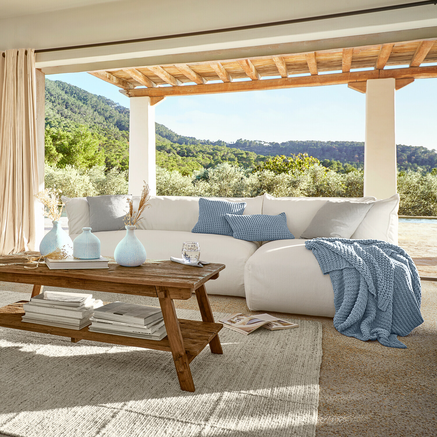 Divano Ysouvie Divano bianco accogliente con coperta e cuscini blu lavorati a maglia su terrazza con vista verde